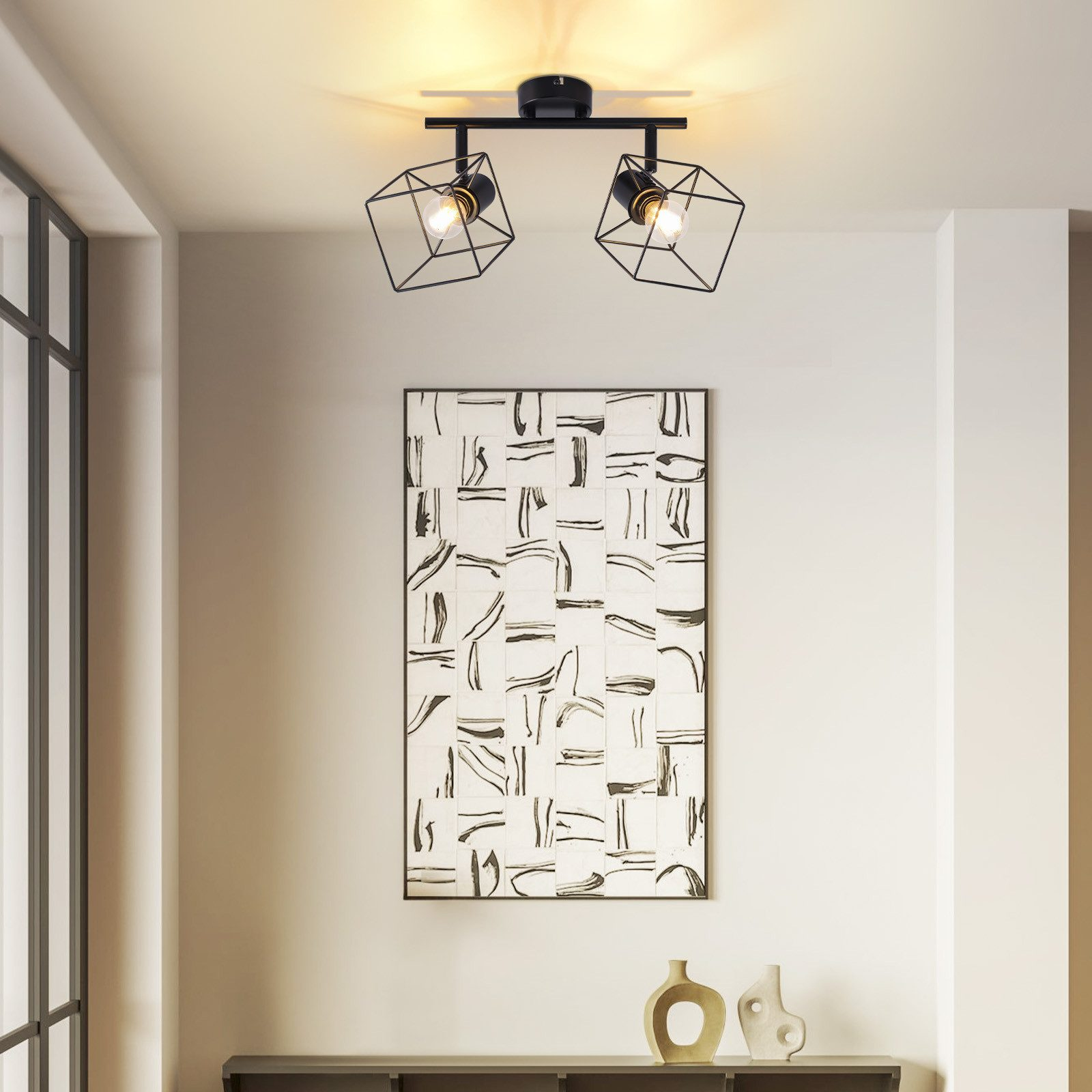 Due faretti neri geometrici montati a soffitto, con un dipinto e vasi sullo sfondo.