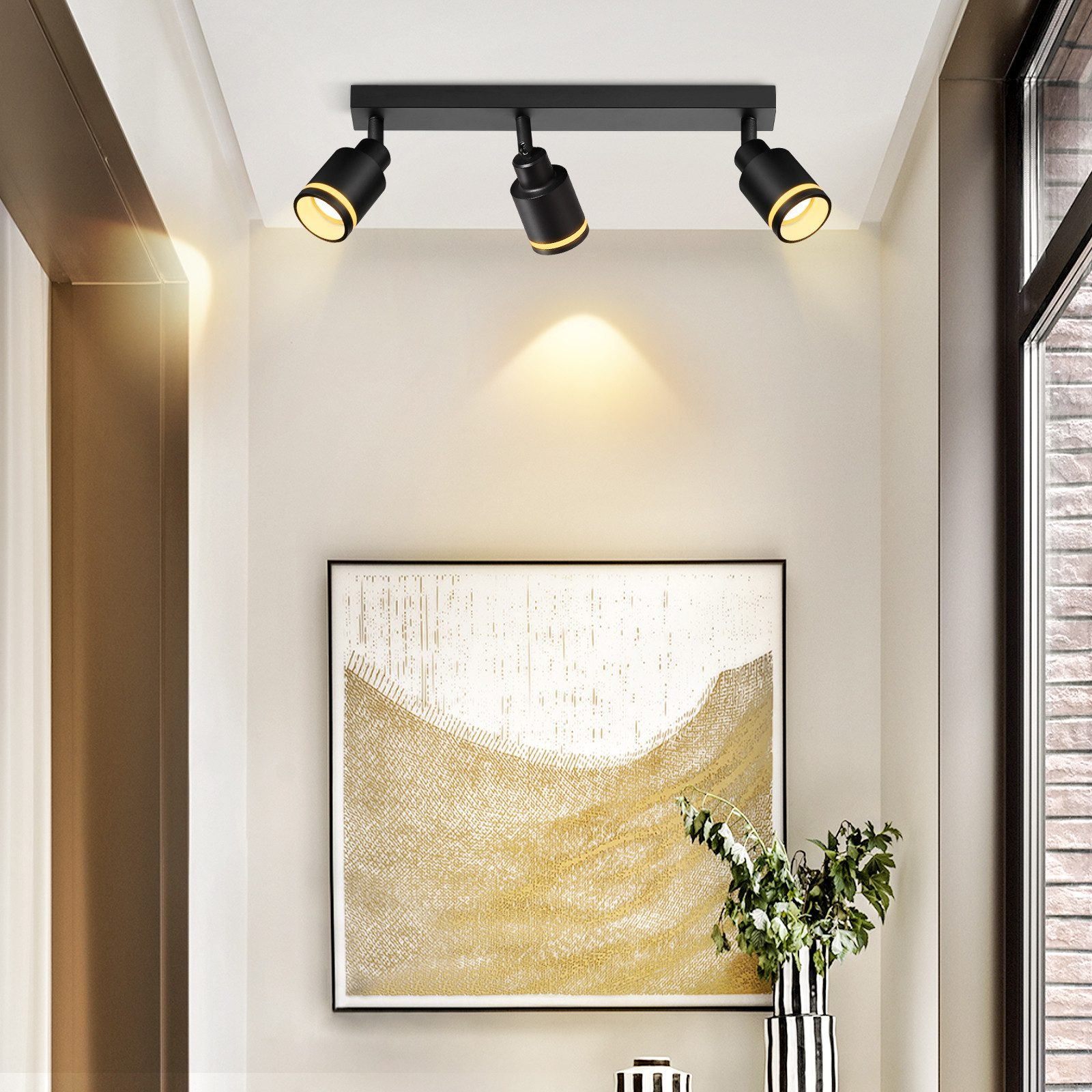Tre faretti neri montati su un soffitto bianco, con un dipinto e un vaso sullo sfondo.