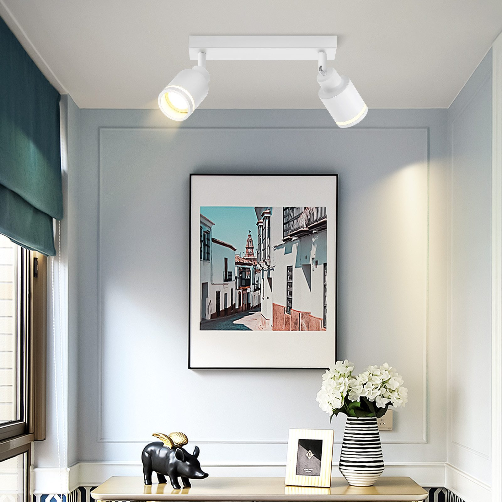 Due faretti bianchi illuminano un quadro e l'arredamento. Sono visibili pareti bianche, tende blu e un vaso.