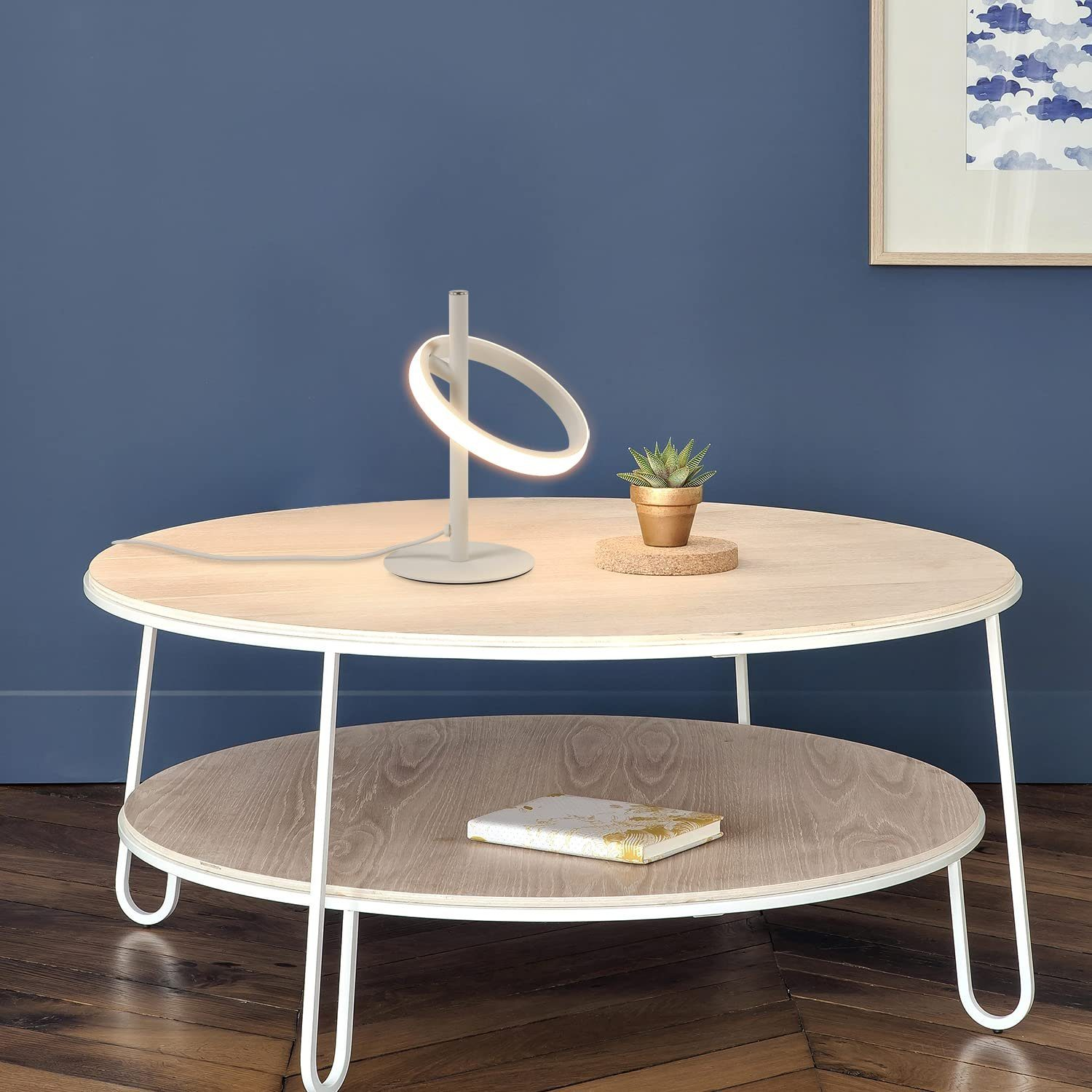 Eine weiße, ringförmige Lampe steht auf einem Holztisch. Eine kleine Pflanze in einem goldenen Topf.