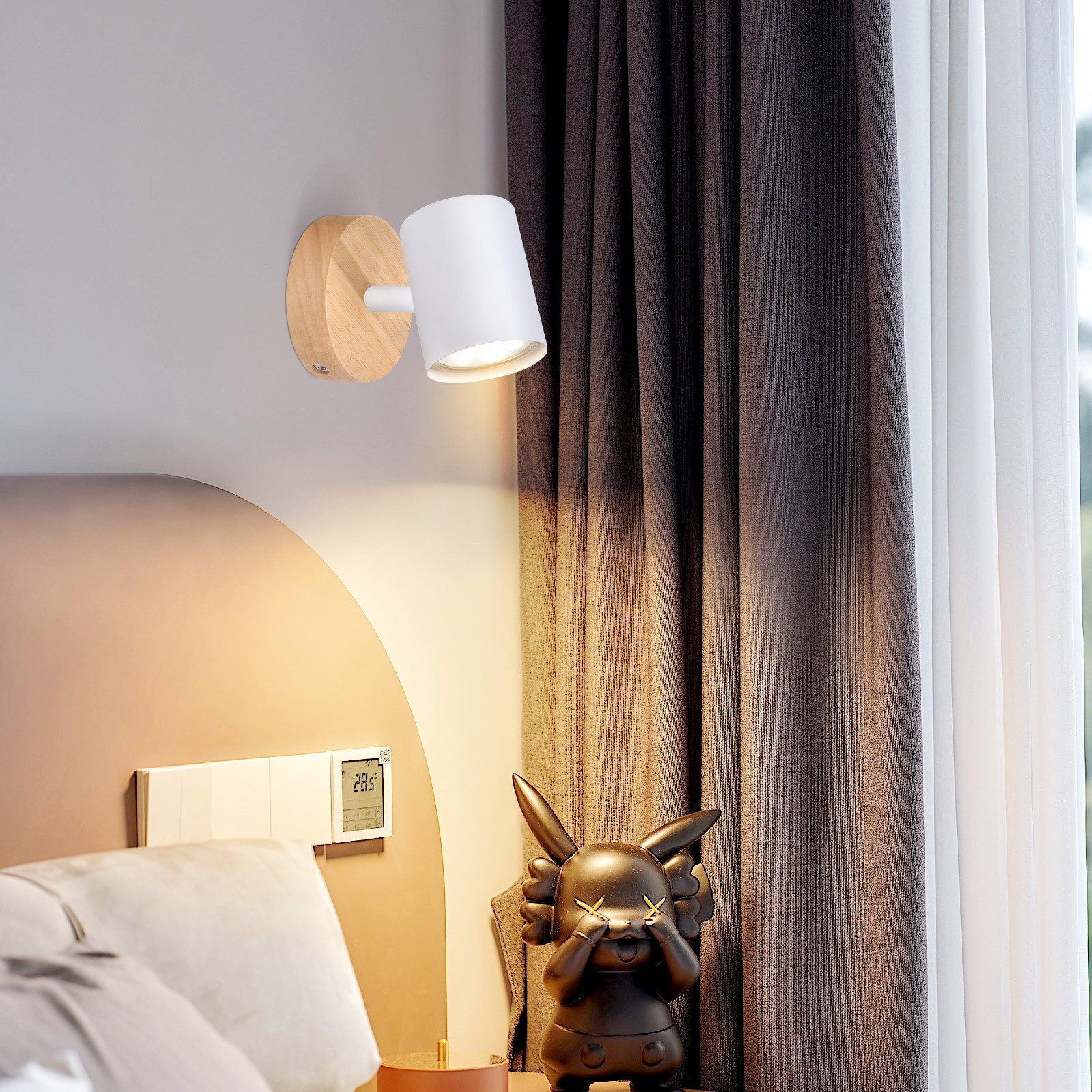 Una lampada da parete con base in legno e paralume bianco, vicino a un letto. Una statuina è vicina.