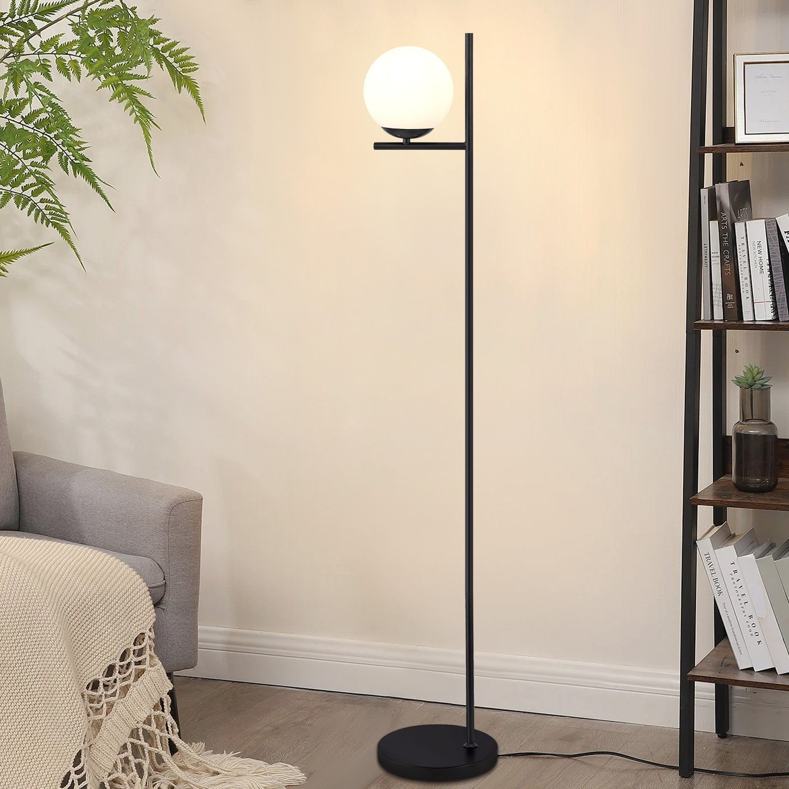 Lampada da terra nera con luce bianca a forma di globo. Si trova accanto a una libreria e a una poltrona grigia.