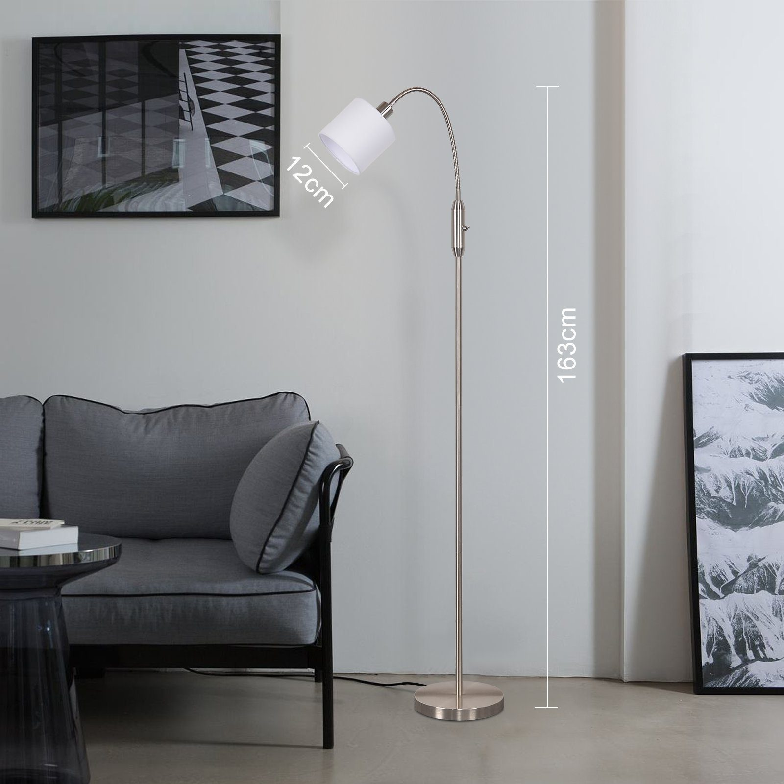 Eine silberne Stehlampe mit weißem Lampenschirm steht neben einem grauen Sofa. Ein Bild hängt an der Wand.