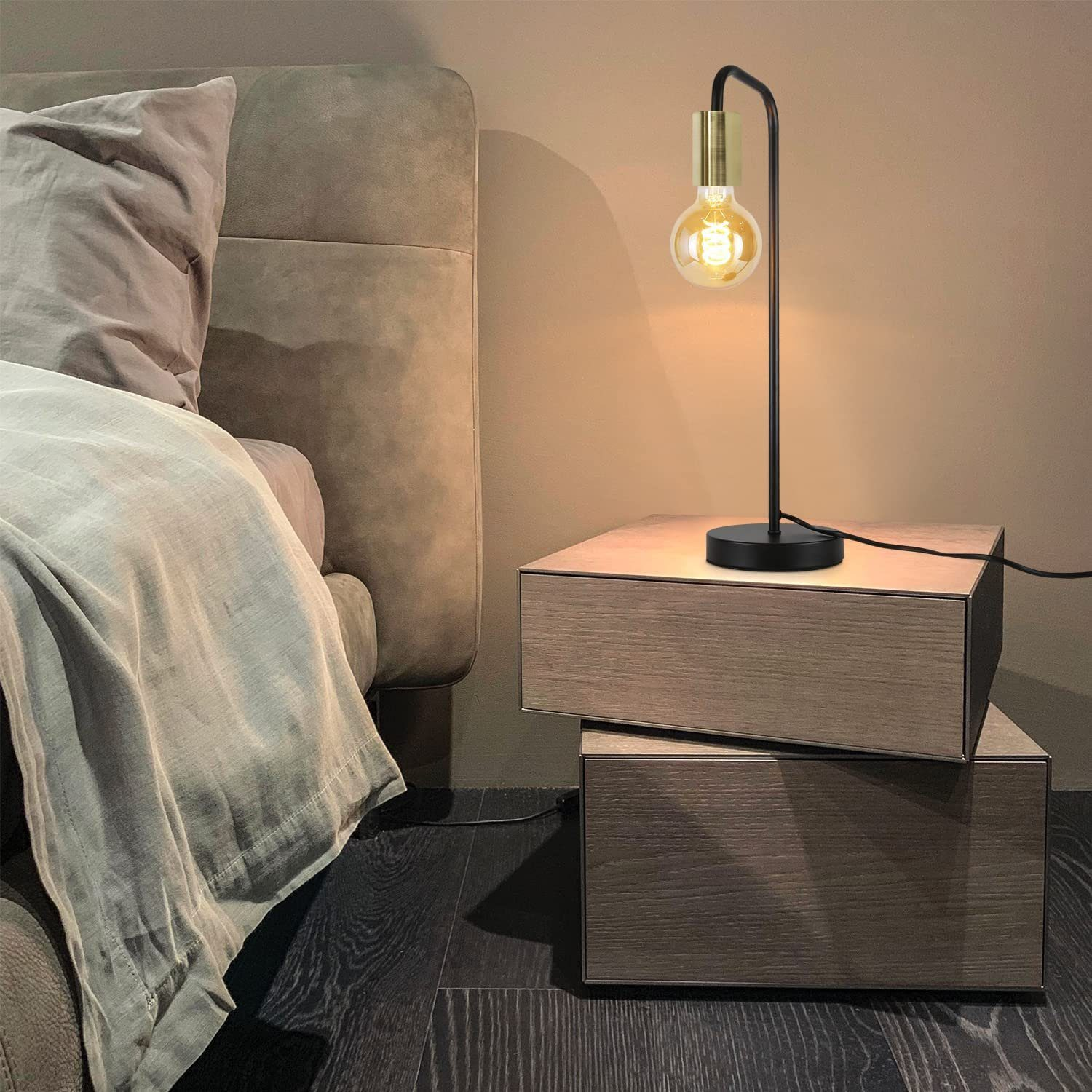 Una lampada da comodino nera e oro con una lampadina visibile, posizionata su un comodino in legno accanto a un letto.