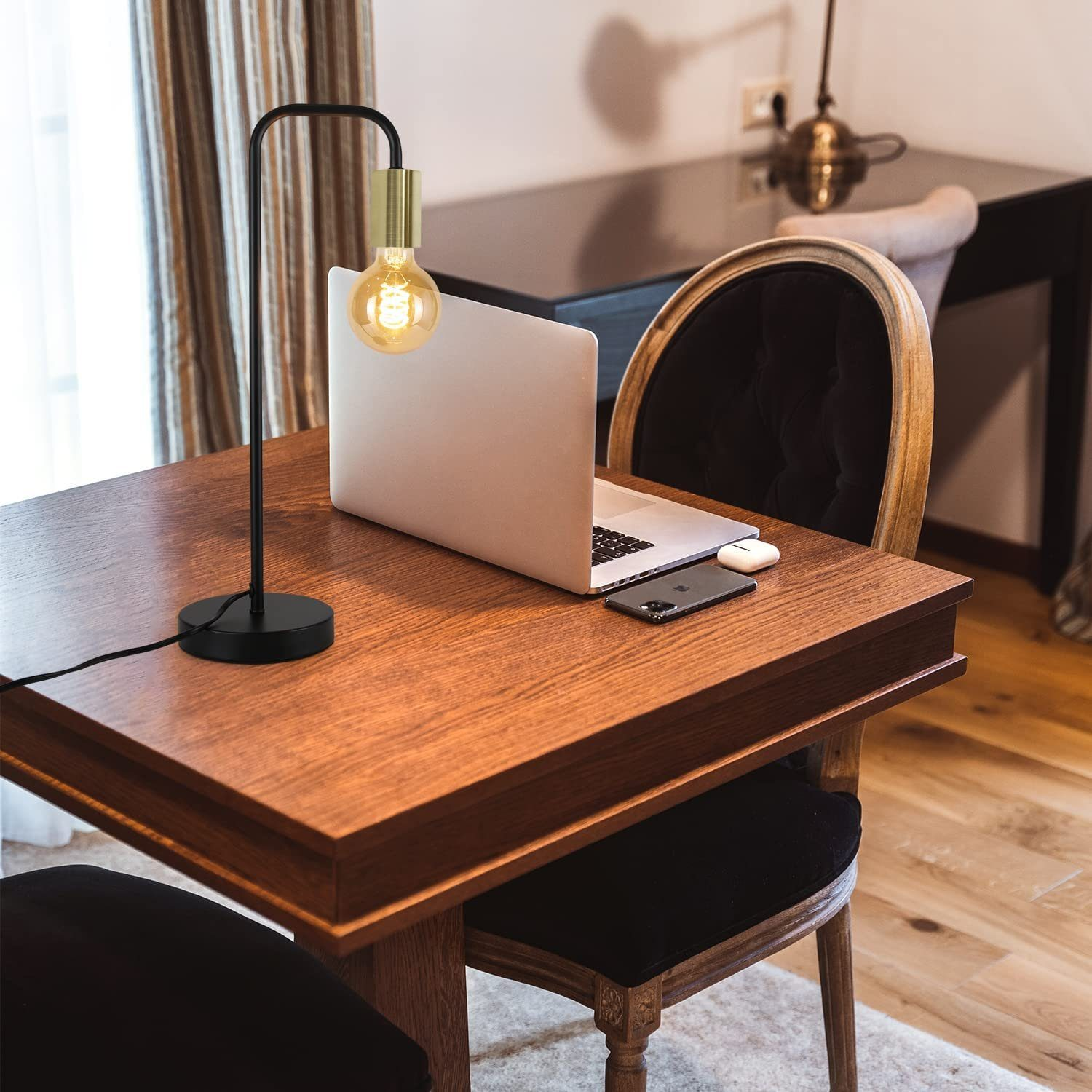 Eine schwarz-goldene Lampe auf einem Holzschreibtisch mit Laptop, Telefon und AirPods.