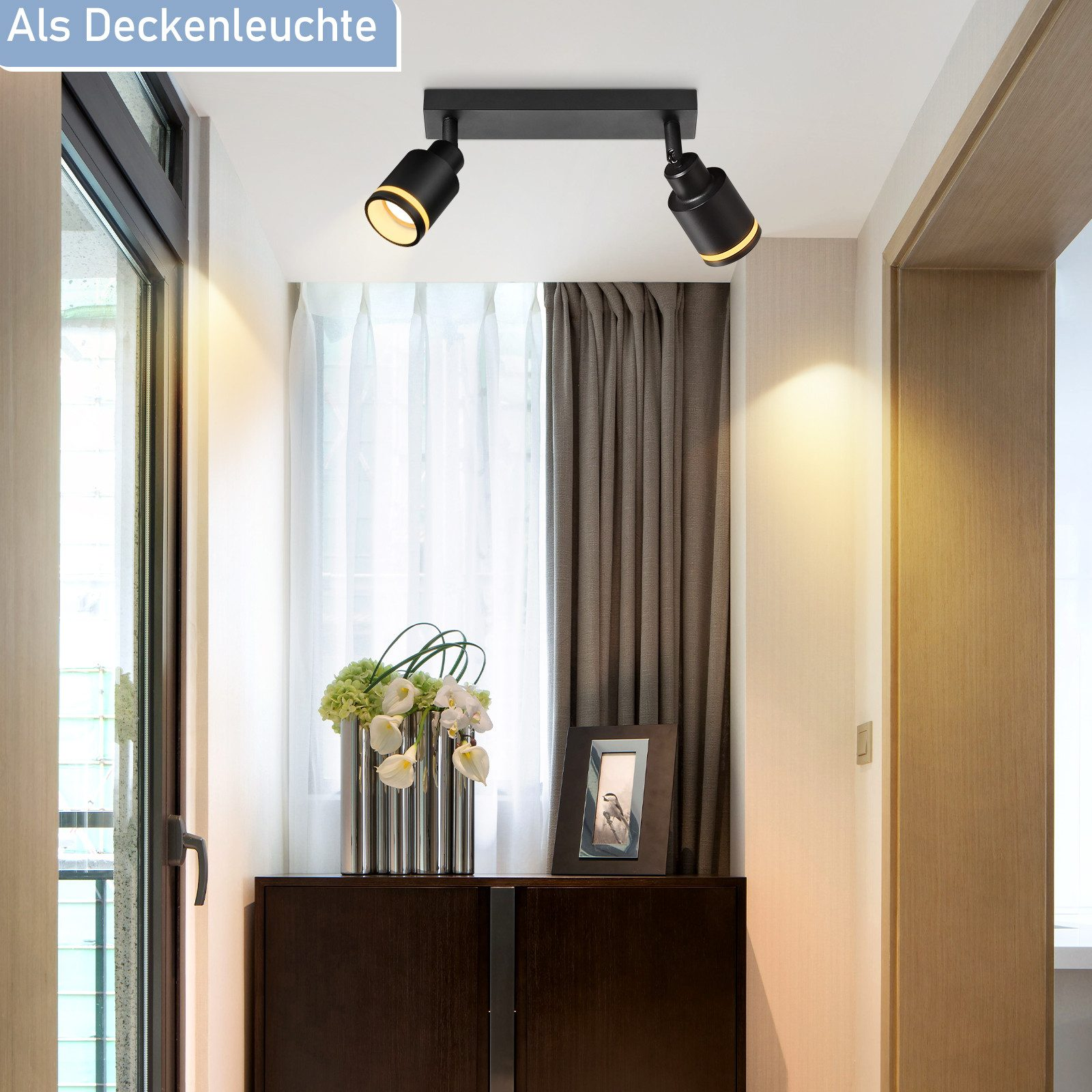 Un corridoio con un'illuminazione a soffitto, fiori in un vaso e una cornice su un mobile.