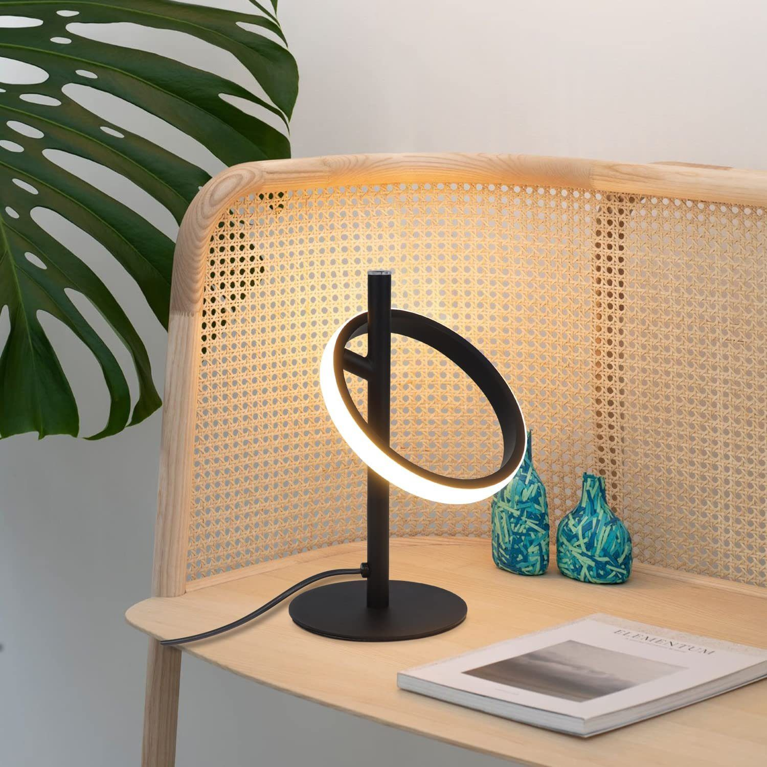 Czarna lampa biurkowa ze światłem pierścieniowym. Stoi na drewnianym biurku obok rośliny i książek.