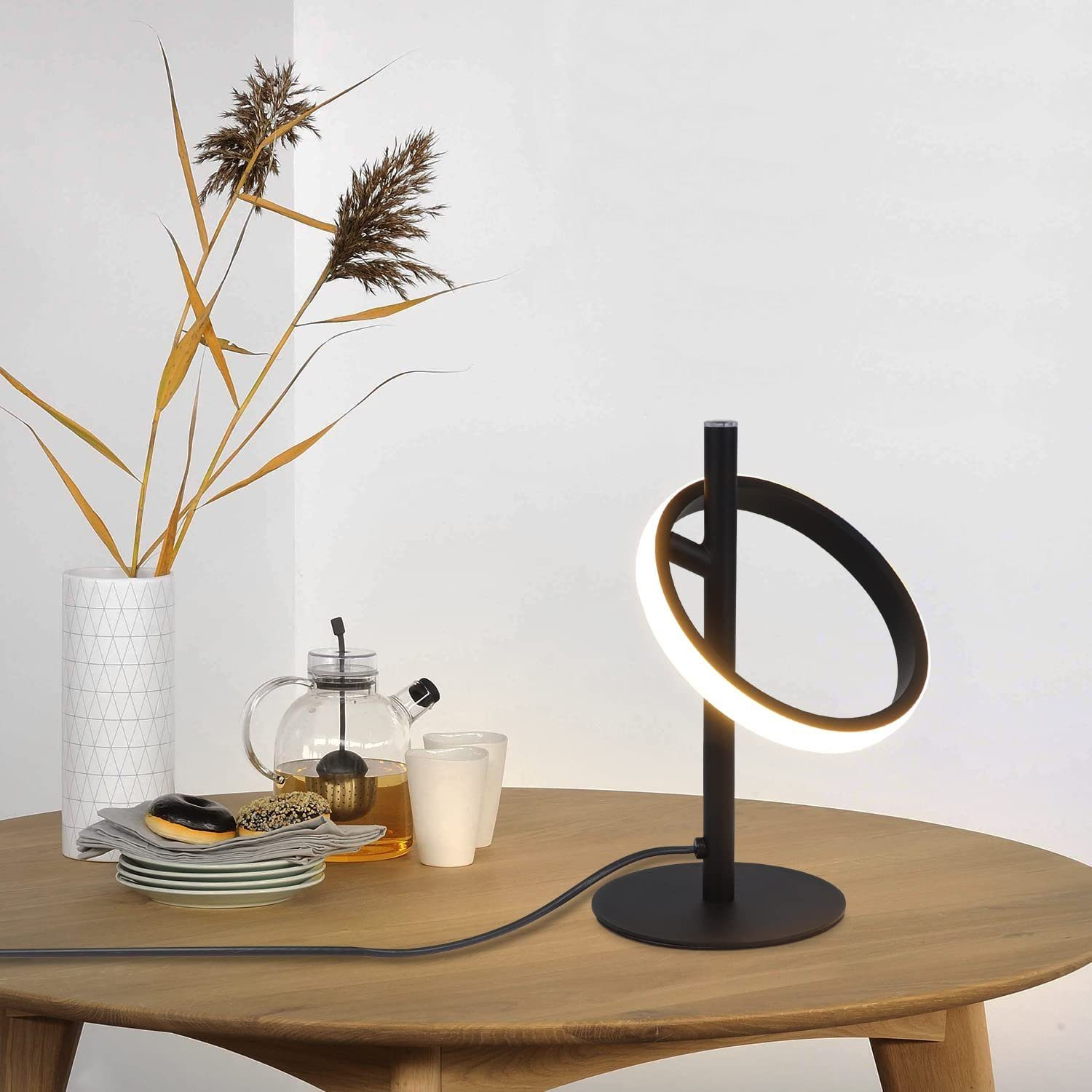 Eine moderne schwarze Tischlampe mit ringförmigem Licht auf einem Holztisch. Teeset und Dekoration sind vorhanden.