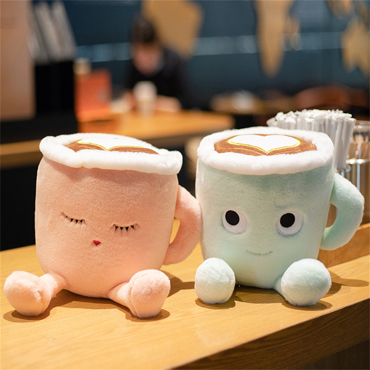 Zwei Plüsch-Kaffeetassen, rosa und mintfarben, mit Herzmustern und geschlossenen Augen, auf einem Holztisch.
