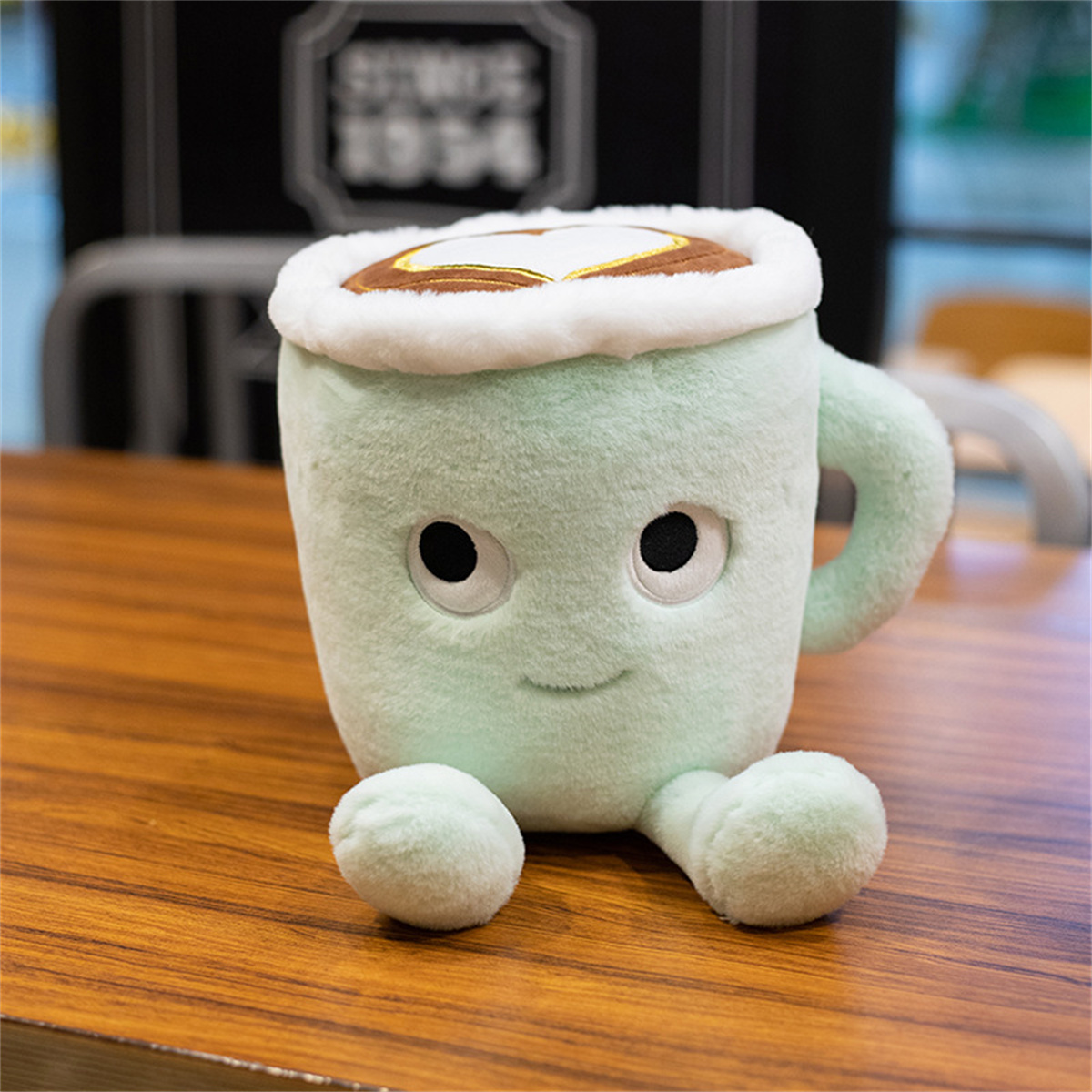 Ein plüschiges, grünes Kaffeetassen-Spielzeug mit einem lächelnden Gesicht. Es hat zwei kleine Füße und einen weißen Schaumstoff.