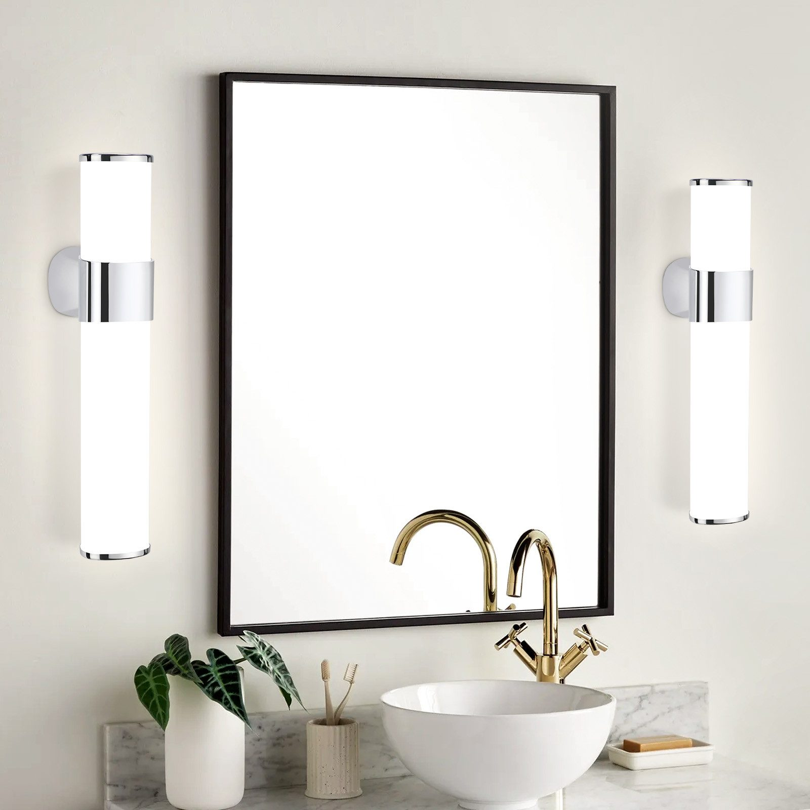 Bagno con specchio, due lampade da parete, lavandino e rubinetto. Pianta sul bordo del lavandino.