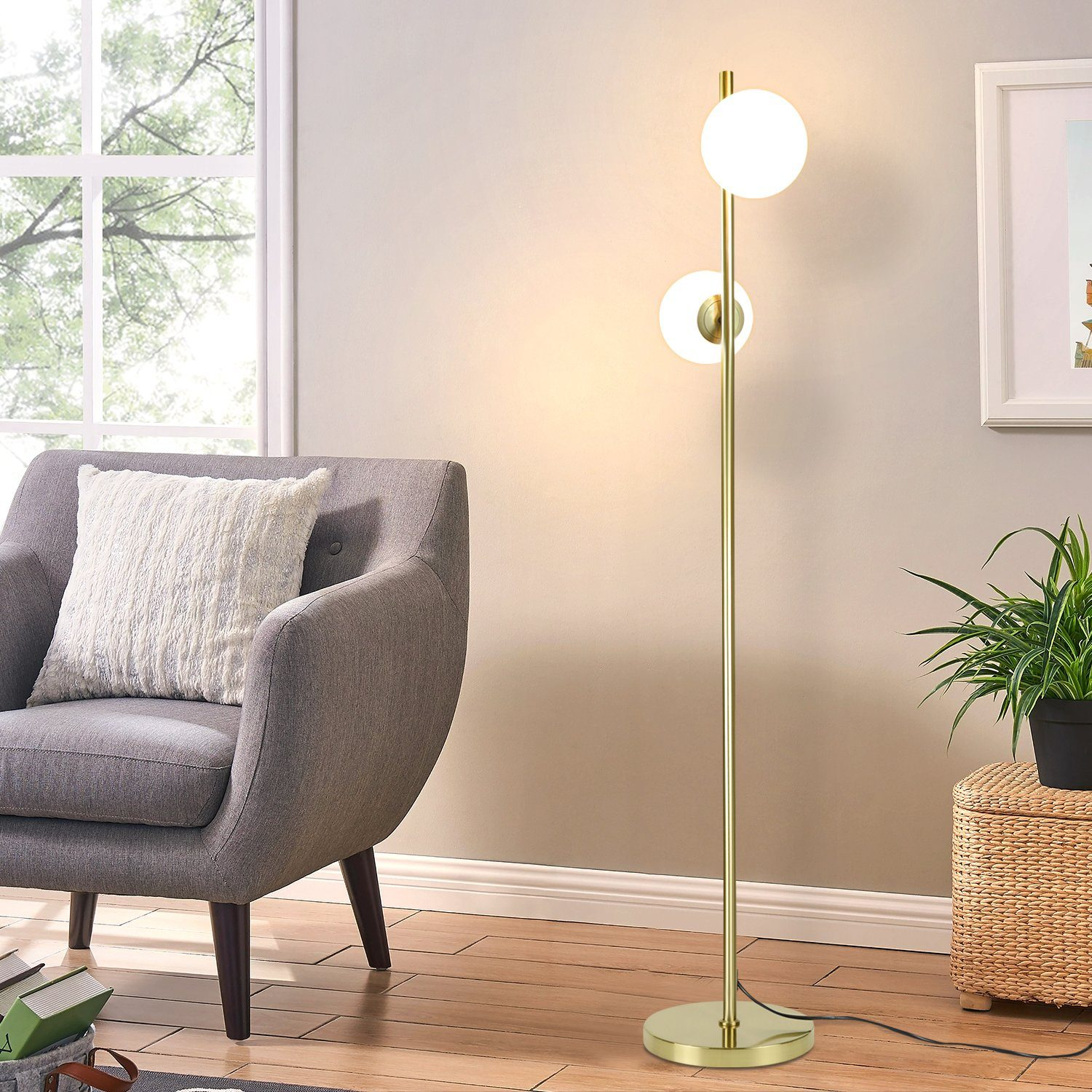 Una lampada da terra dorata con due globi bianchi. Si trova accanto a una poltrona grigia e a un cesto intrecciato.
