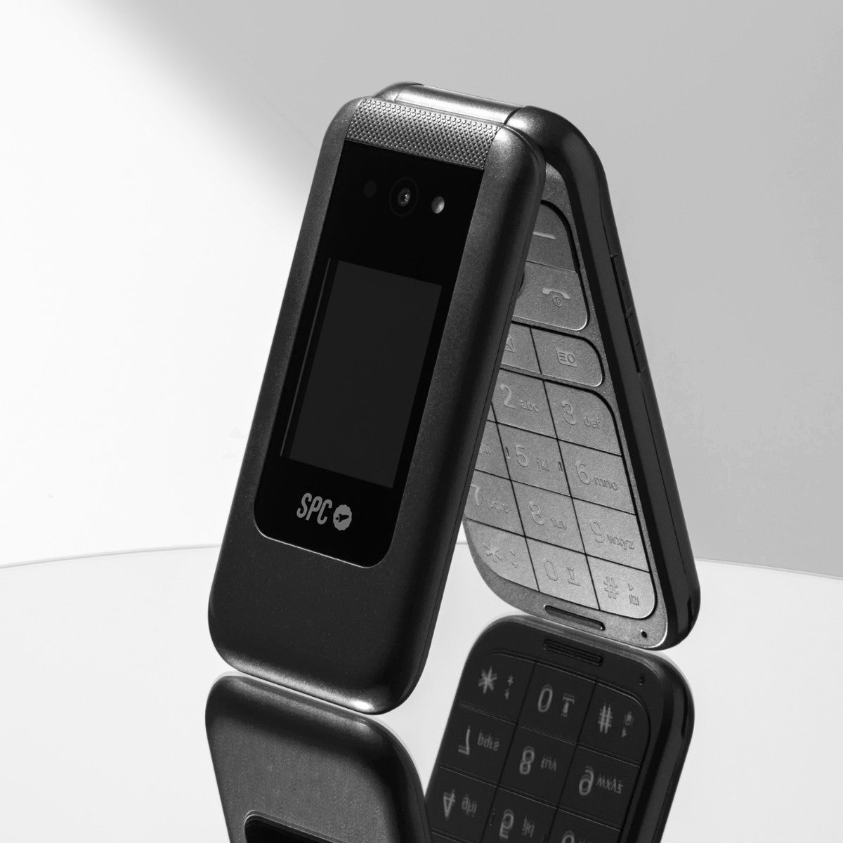 Czarny telefon z klapką otwarty, pokazujący klawiaturę i ekran, na odblaskowej powierzchni.