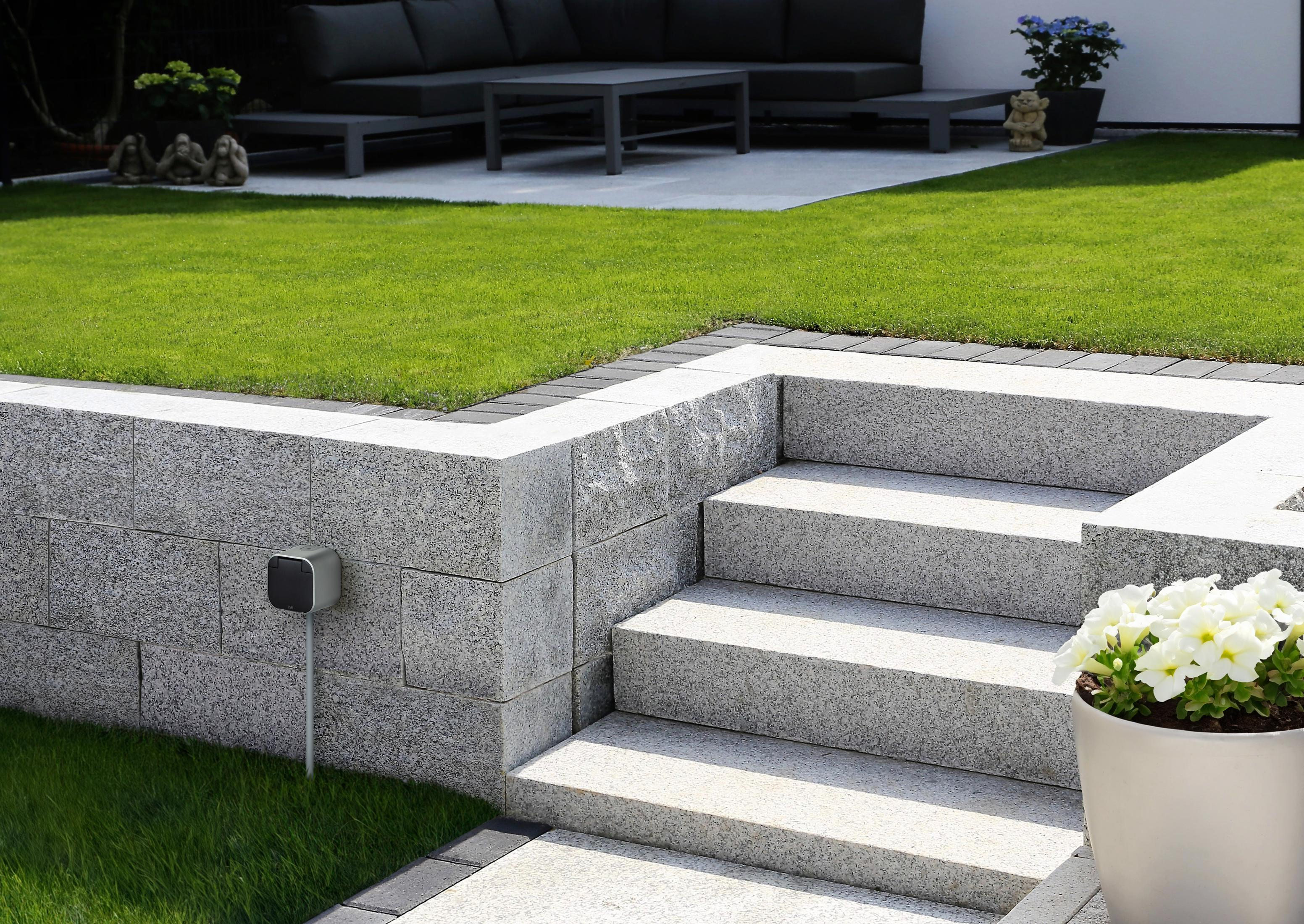 Steintreppe in einem Garten, neben einer Mauer. Grünes Gras und ein Blumentopf sind sichtbar.