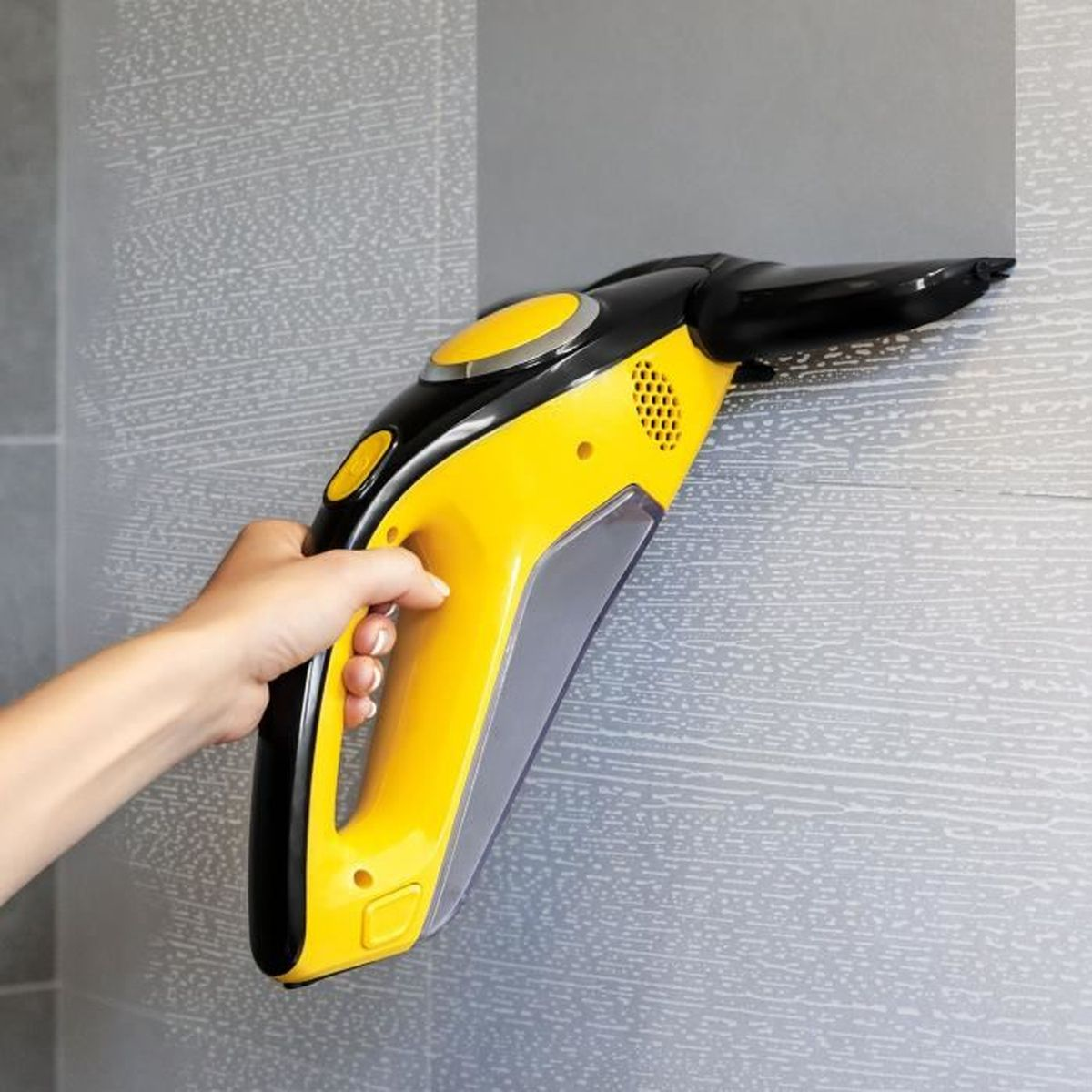 Person verwendet einen gelb-schwarzen Fensterreiniger an einer grau gefliesten Wand. Hand sichtbar.