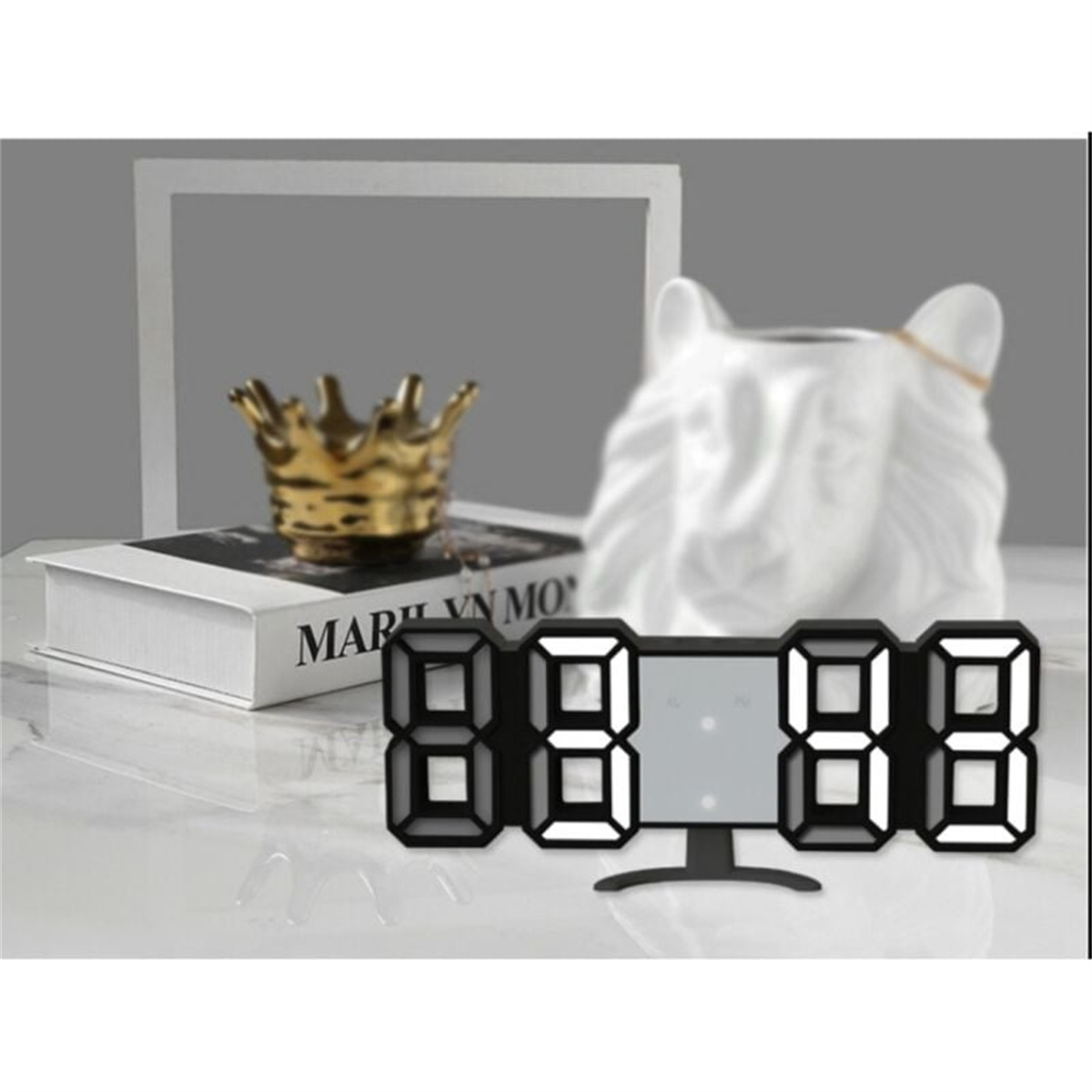 Eine Digitaluhr mit einer Krone, einem Buch und einer Keramikkatze. Die Uhr zeigt die Zeit an.