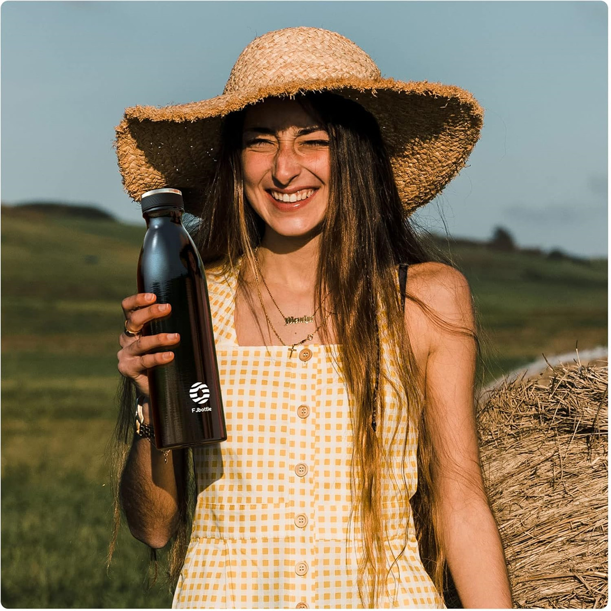 Eine lächelnde Frau mit Strohhut, die eine schwarze Wasserflasche hält, an einem sonnigen Tag.