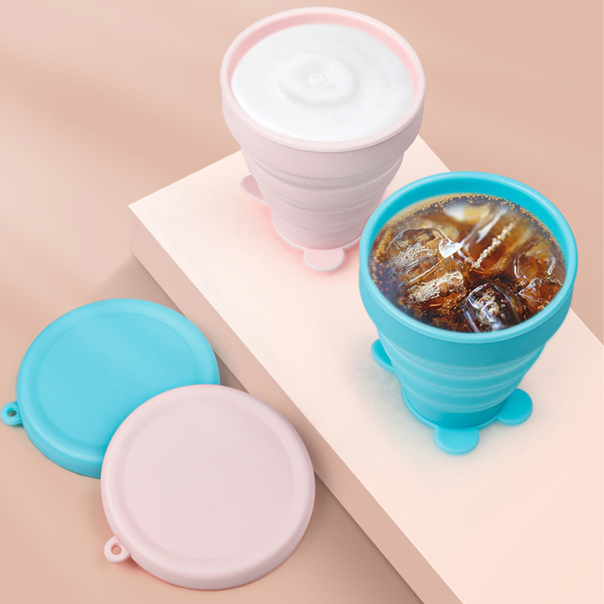 Rosa und blaue zusammenklappbare Becher mit Deckeln, auf rosa Oberfläche. Ein Becher hat Eis und Flüssigkeit.