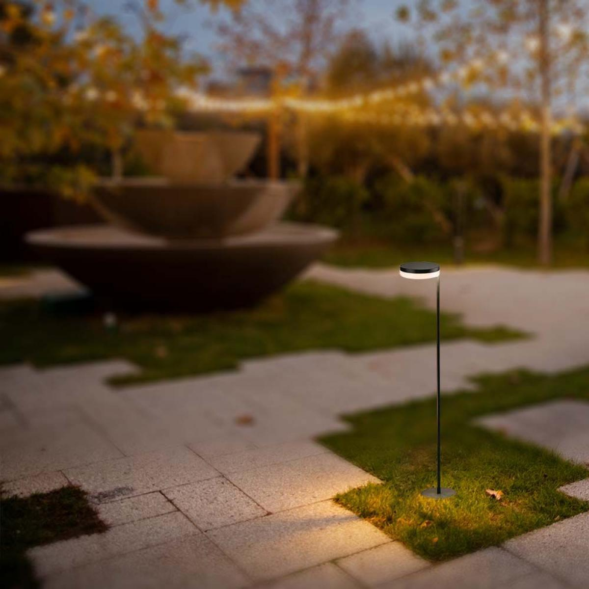 Un lampione nero illumina un sentiero di pietra e l'erba in un giardino sfocato. Una fontana è sullo sfondo.