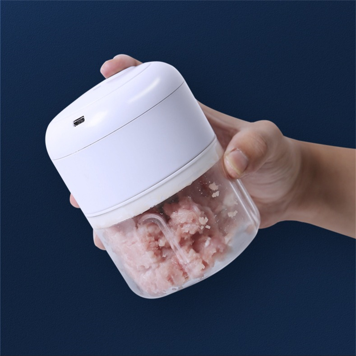Ein weißer Mini-Zerkleinerer mit Fleisch darin. Von einer Hand gehalten, auf blauem Hintergrund.