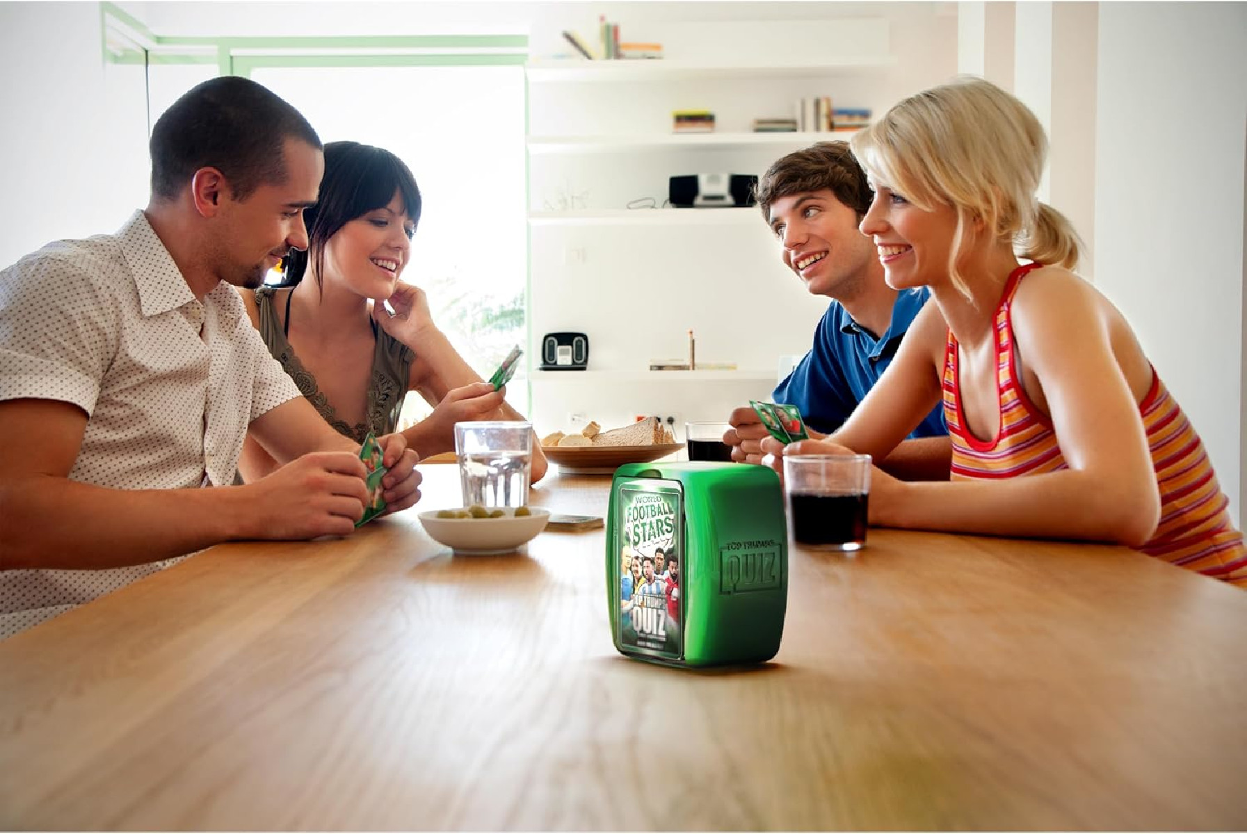 Vier Personen spielen ein Brettspiel. Eine grüne Box ist sichtbar. Hintergrund: Raum mit Holztisch.