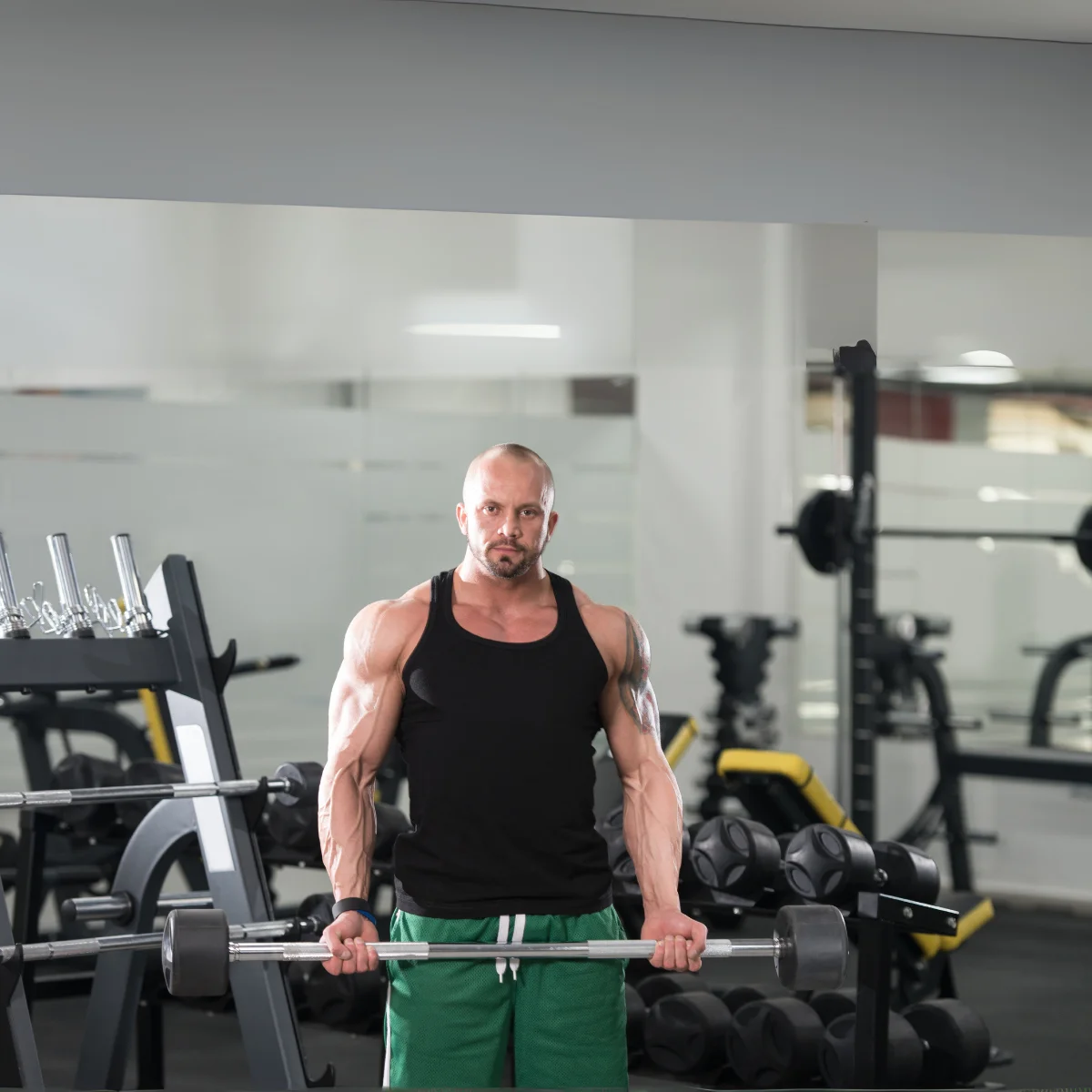 Mann im Fitnessstudio, der eine Langhantel hebt, ein schwarzes Tanktop und grüne Shorts trägt.