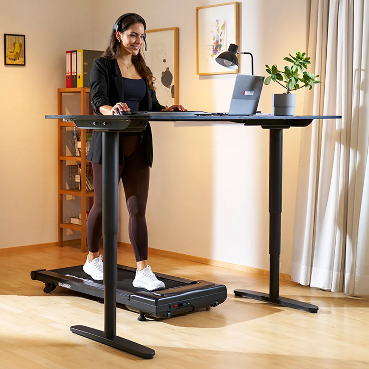 Frau arbeitet am Laptop, während sie an einem Laufband-Schreibtisch steht. Pflanze und Lampe stehen auf dem Schreibtisch.