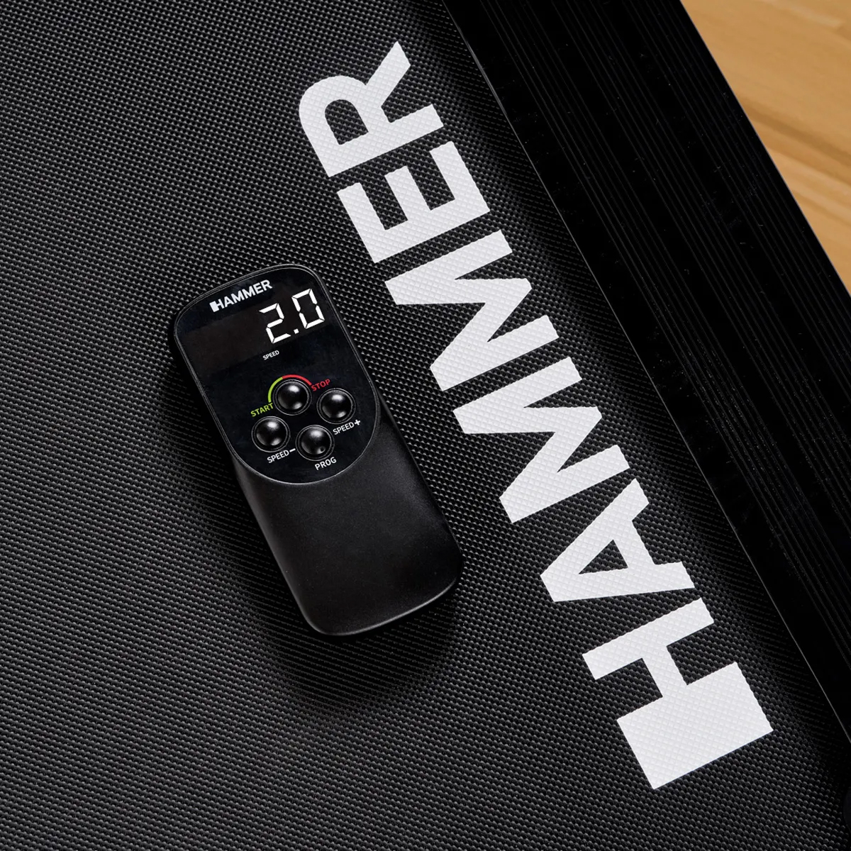 Schwarze Fernbedienung auf einer schwarzen Laufbandoberfläche. Hammer-Logo und Geschwindigkeitseinstellungen sichtbar.