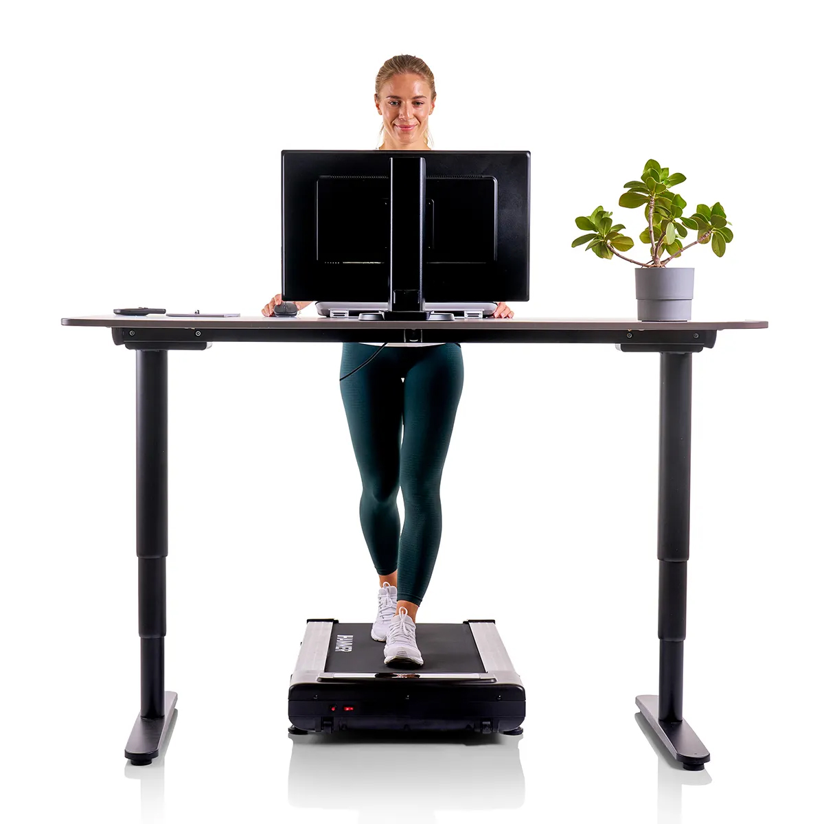 Frau auf Laufband hinter einem Schreibtisch mit Monitoren und einer Pflanze.