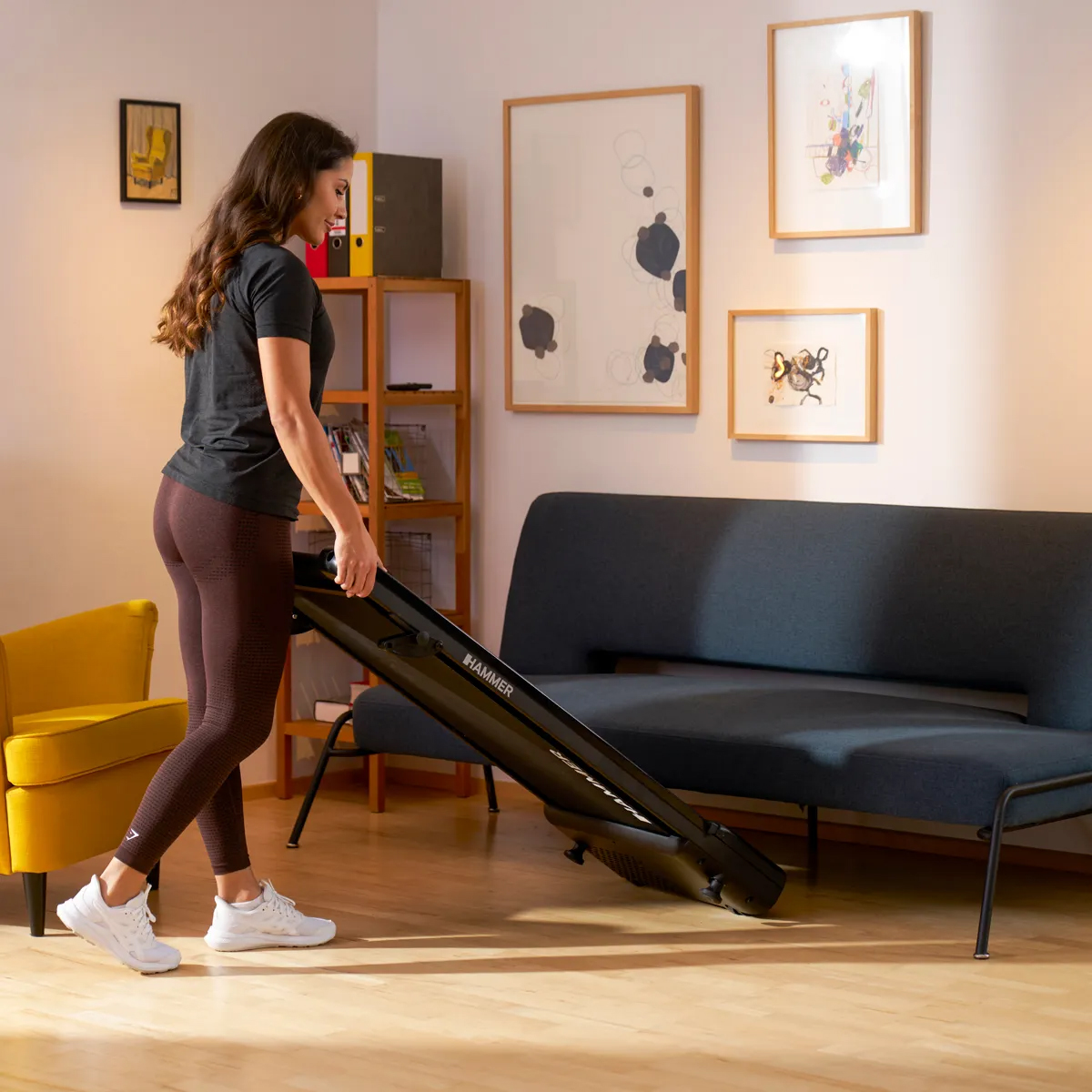 Eine Frau in Leggings trägt ein schwarzes Trainingsgerät in einem Wohnzimmer mit Kunstwerken und Möbeln.