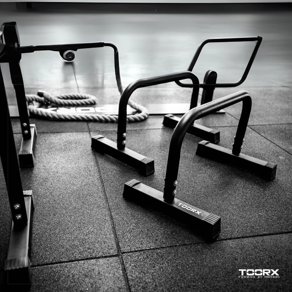 Black and white gym scene. Parallel bars, weights, and a rope are present in the image.