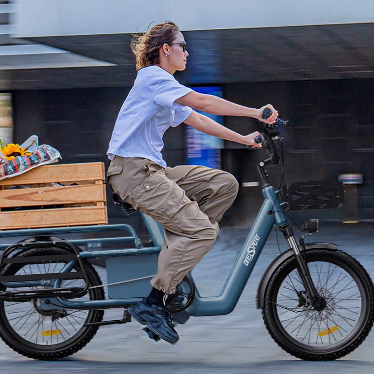 Osoba jadąca rowerem cargo elektrycznym. Rower ma drewnianą skrzynię ładunkową i czarny przedni kosz. Osoba jest w ruchu.