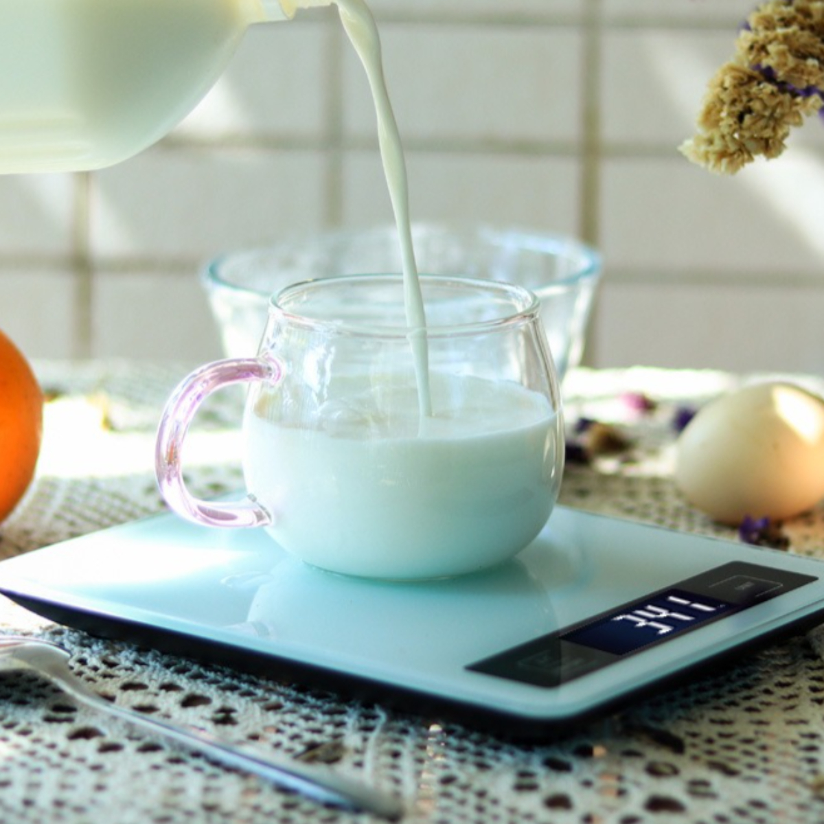 Milch wird in eine Glas-Tasse auf einer Waage gegossen. Die Tasse hat einen pinken Griff. Andere Gegenstände im Hintergrund.
