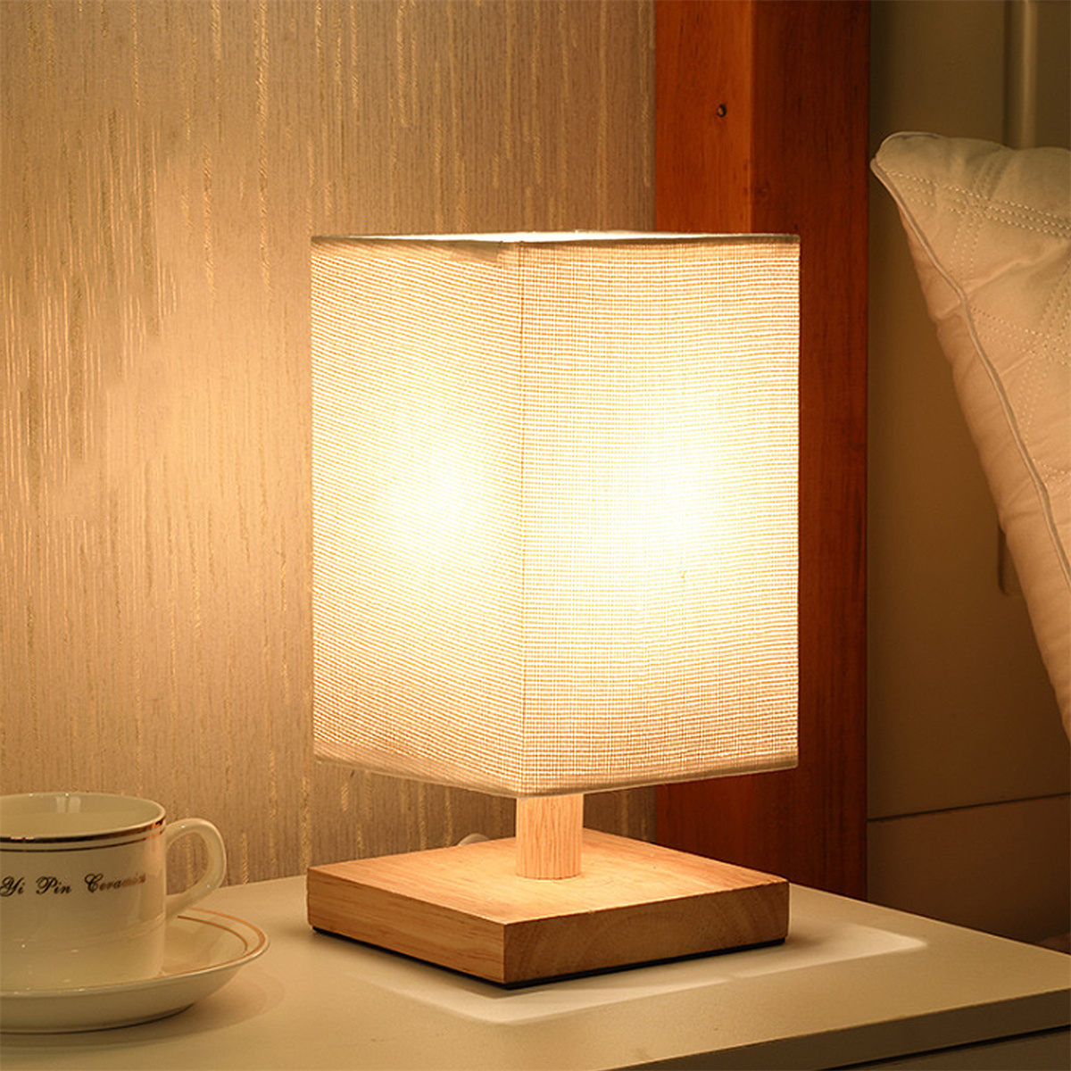 Eine beleuchtete Tischlampe auf Holzsockel, neben einer Tasse und Untertasse auf einem Nachttisch.