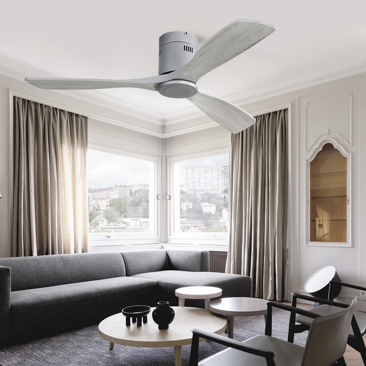 Ein Deckenventilator mit drei hellgrauen Flügeln. Es befindet sich in einem Raum mit Sofa und Vorhängen.