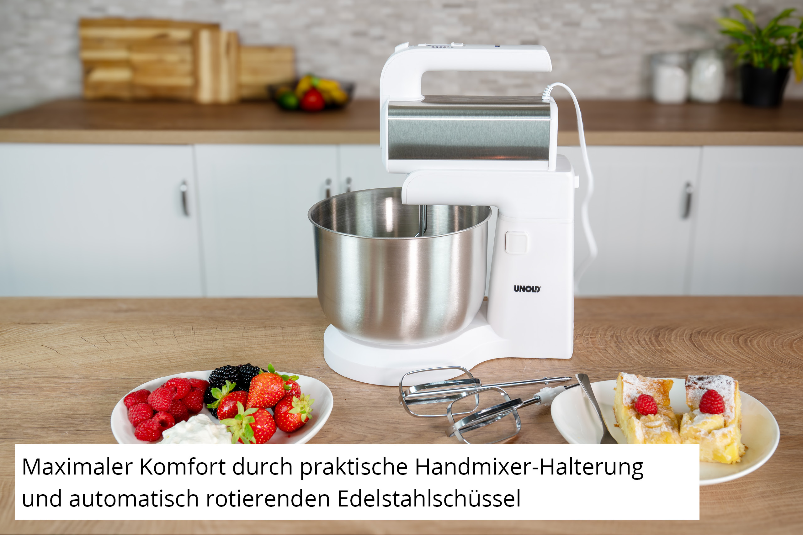 Ein weißer Küchenmixer mit silberner Schüssel und Rührbesen auf einem Holztisch. Beeren und Kuchen sind ebenfalls auf dem Tisch.