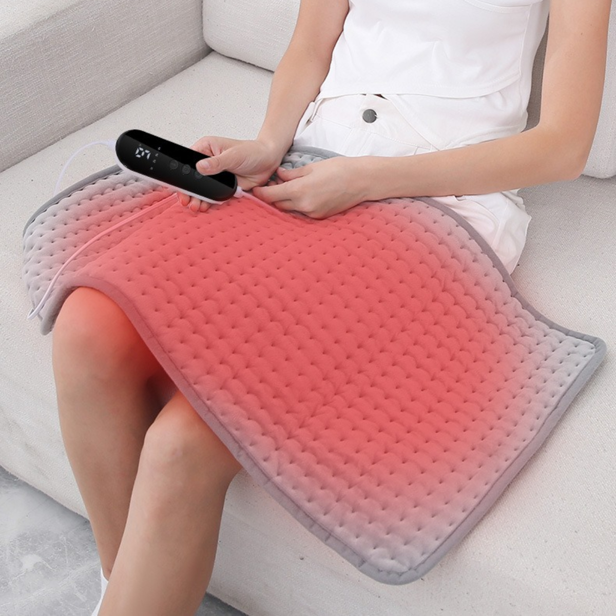 Eine Person sitzt auf einem Sofa und benutzt ein Heizkissen, das sie mit einem schwarzen Gerät steuert.