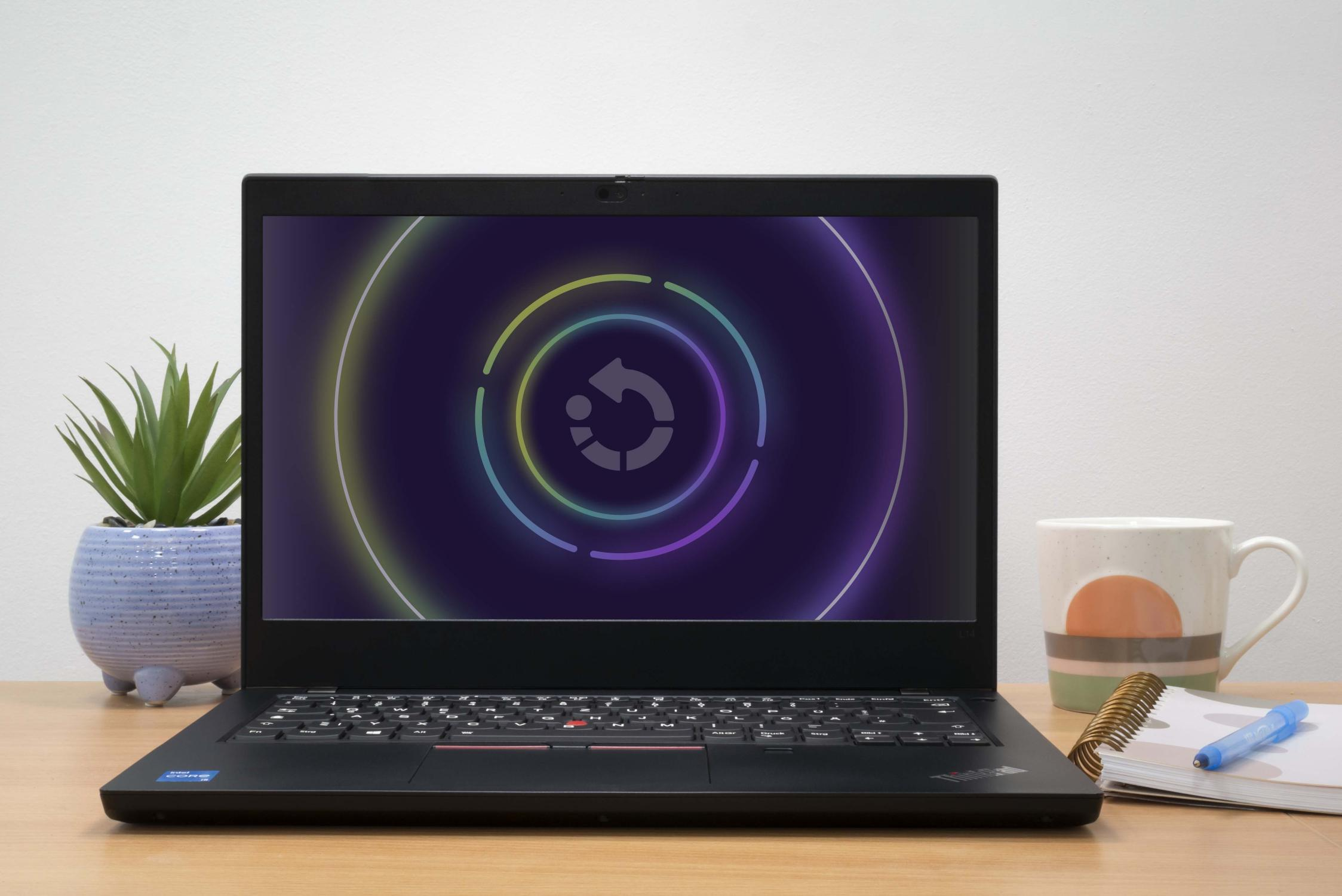 Laptop auf einem Holzschreibtisch, zeigt einen bunten Bildschirm. Eine Pflanze und eine Tasse sind auch da.