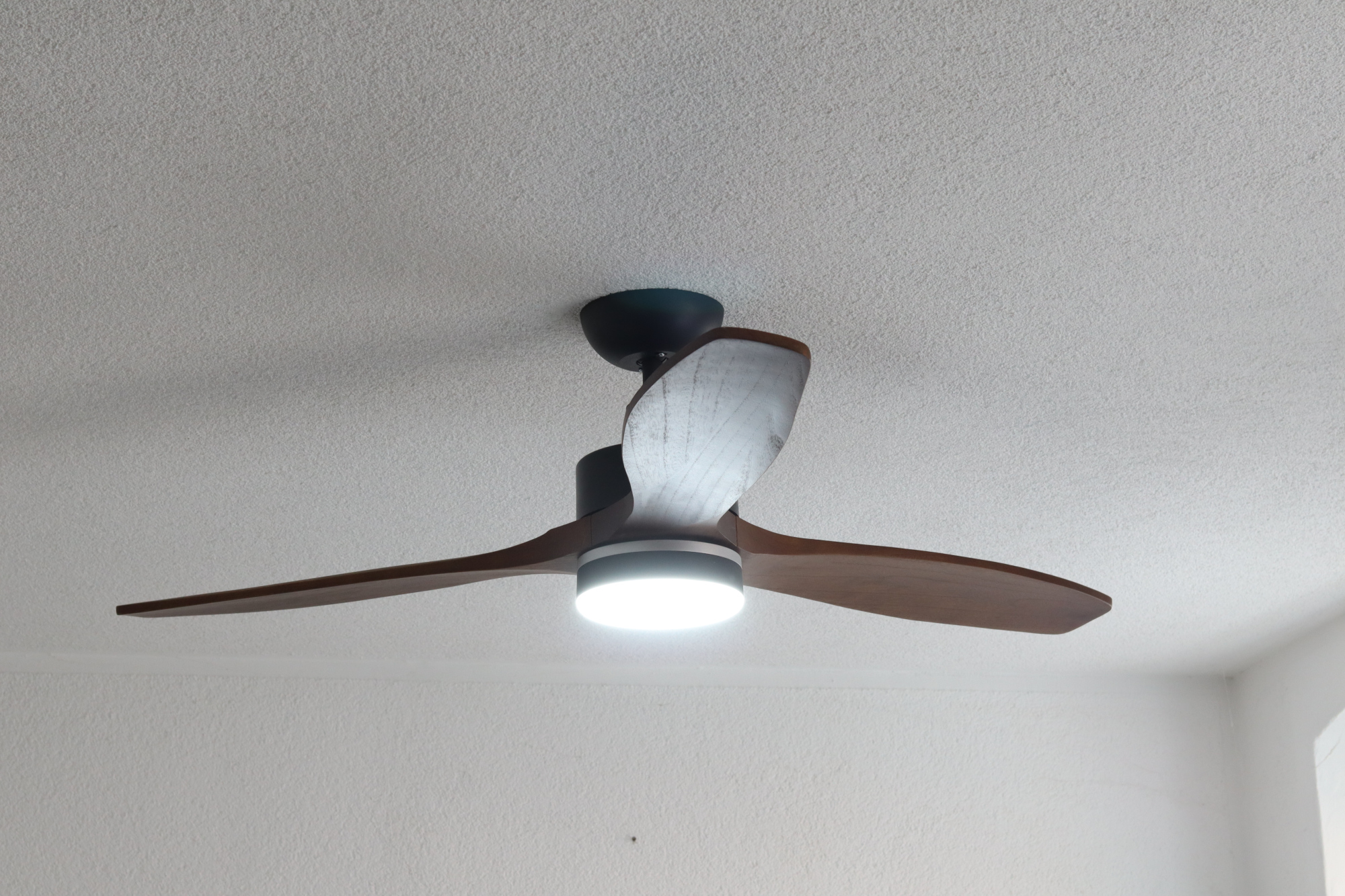 Ein Deckenventilator mit Holzflügeln und einer zentralen Leuchte. Die Decke ist weiß.