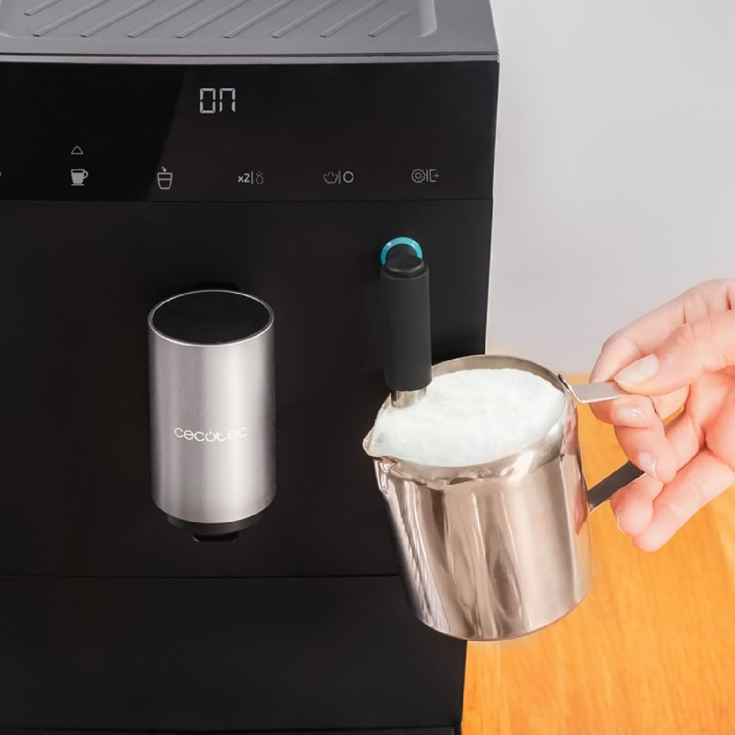 Una mano versa schiuma di latte in una caraffa in acciaio inox. Una macchina da caffè nera sullo sfondo.
