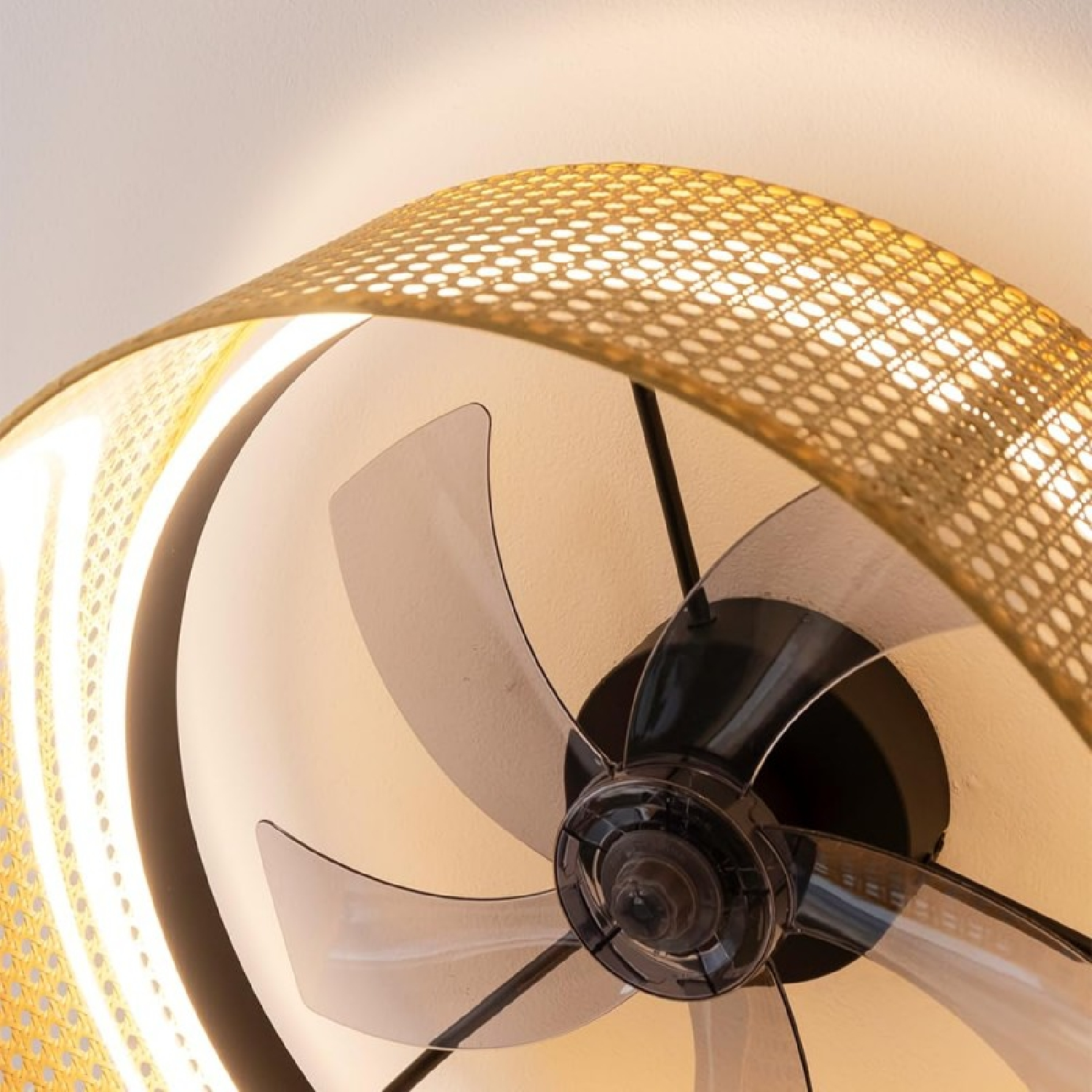 Primer plano de un ventilador con aspas, con pantalla tejida. El ventilador está en el techo blanco.