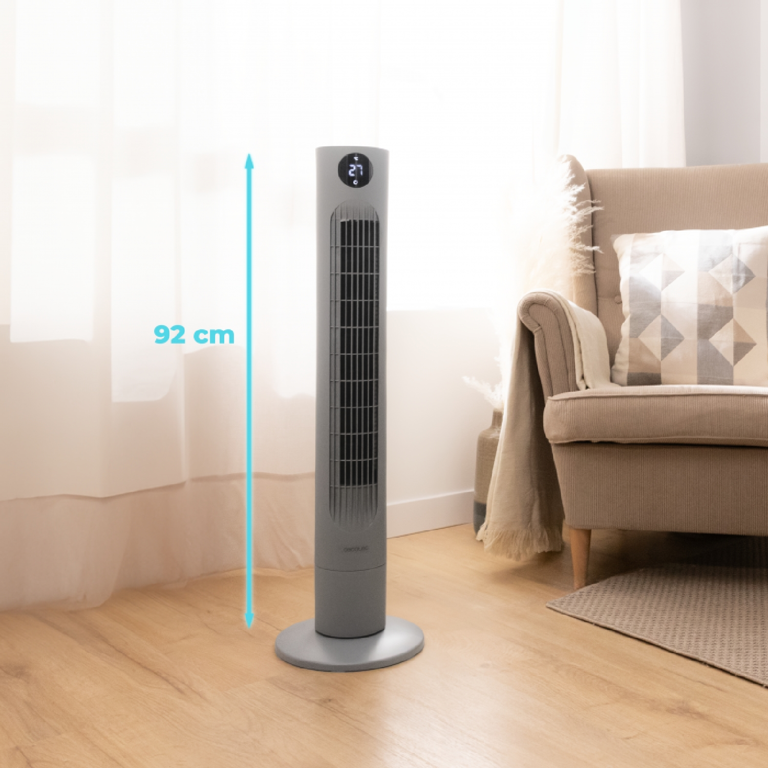 Un ventilador de torre gris, de 92 cm de altura, junto a un sillón beige en una habitación con suelo de madera.