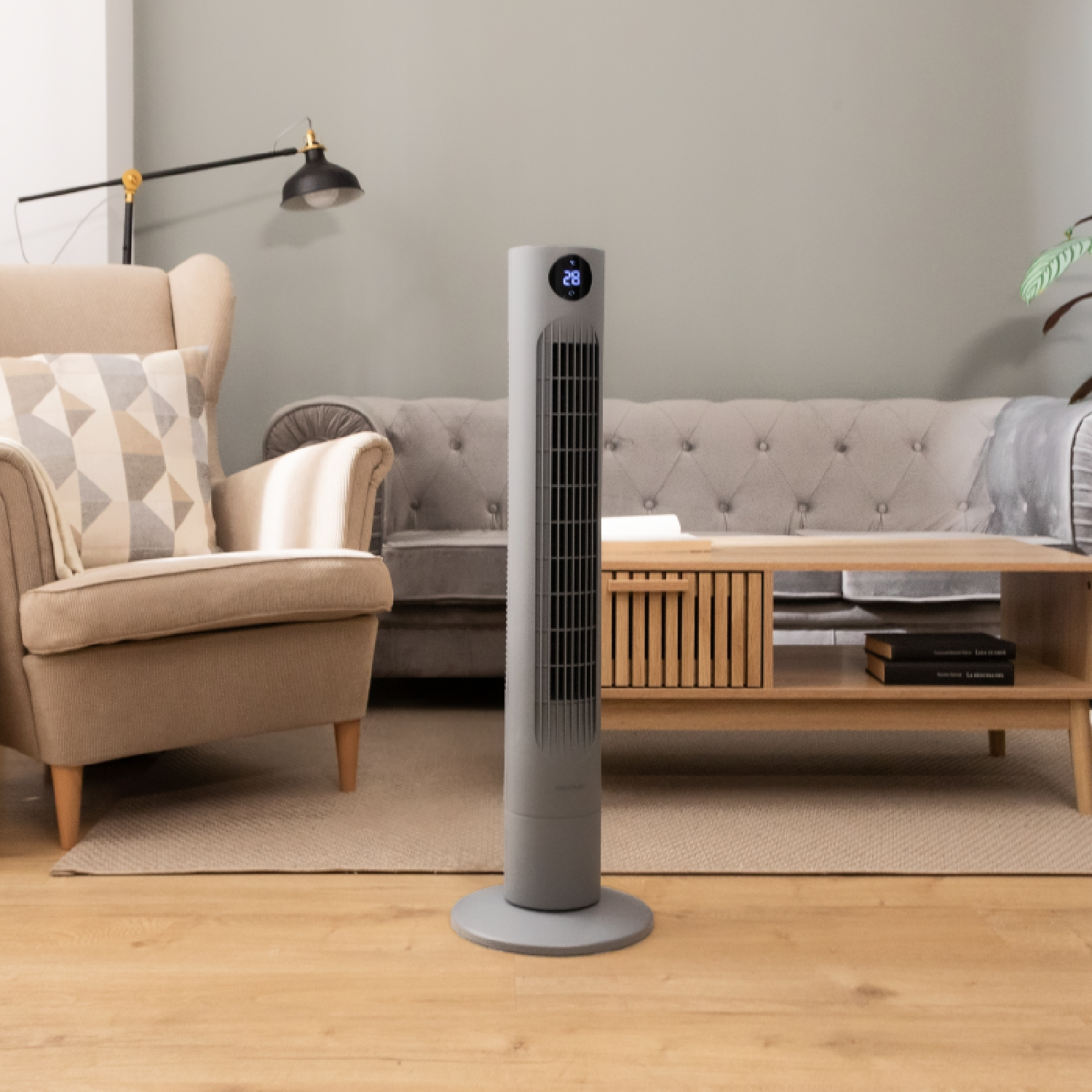 Ventilador de torre gris en salón. Sofá beige, mesa de centro de madera y planta verde. Paredes grises.