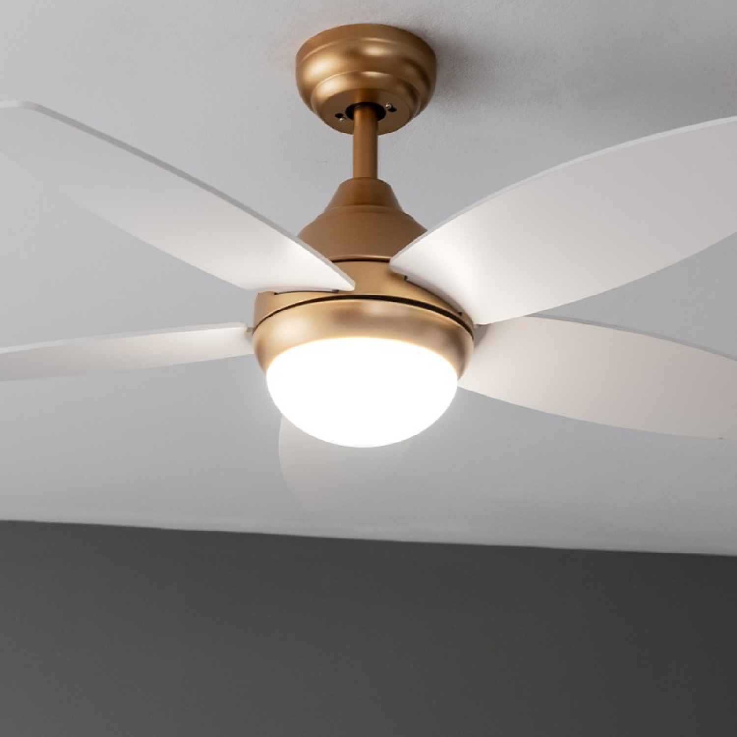 Deckenventilator mit goldener Halterung, weißen Flügeln und zentralem Licht an weißer Decke.