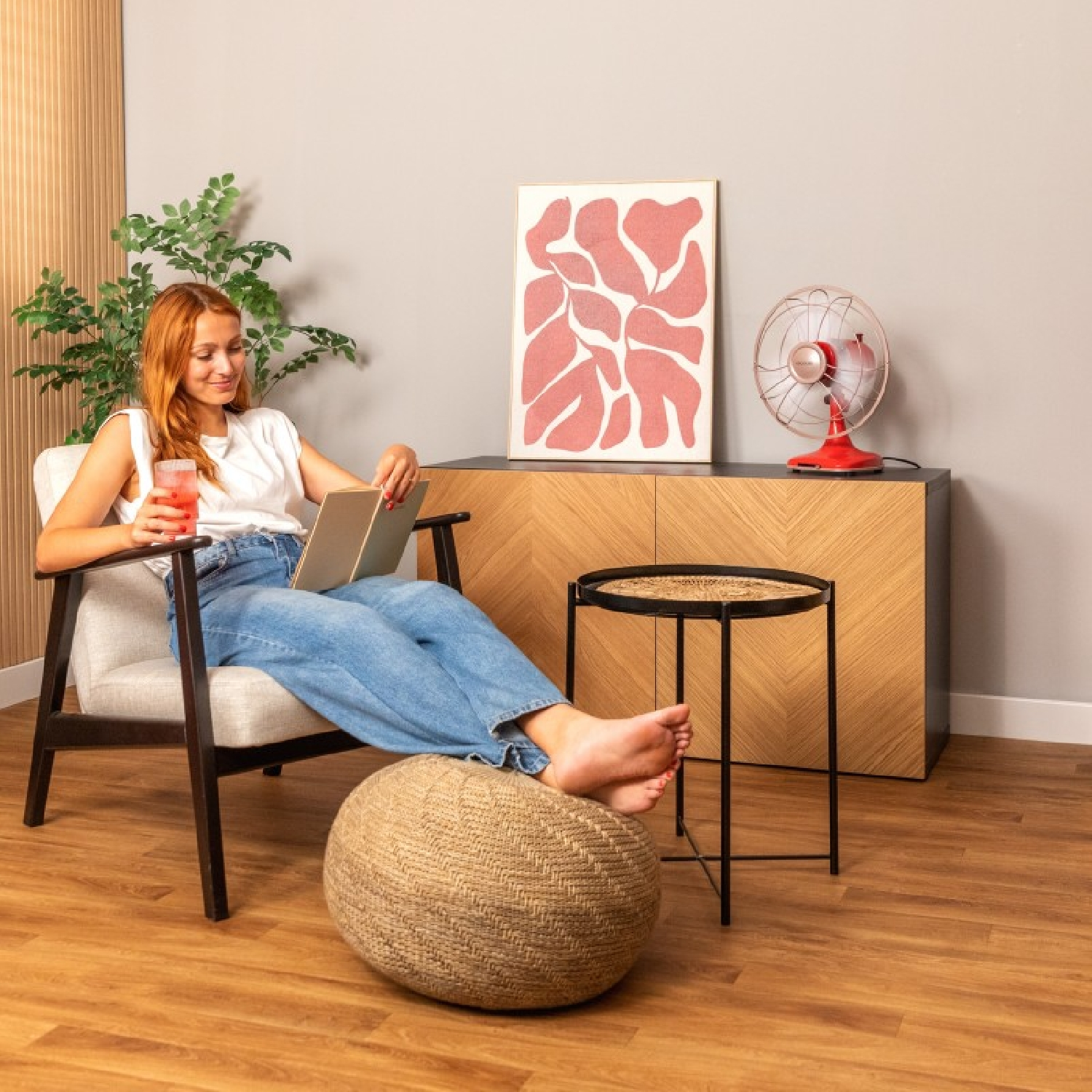 Una donna seduta su una sedia, legge. Un tavolino e un ventilatore sono vicini. Un quadro è appeso al muro.