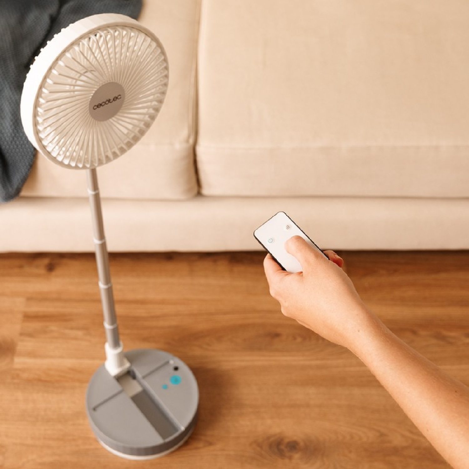 Osoba trzyma pilota. Biały, składany wentylator na podłodze. Neutralne tło.
