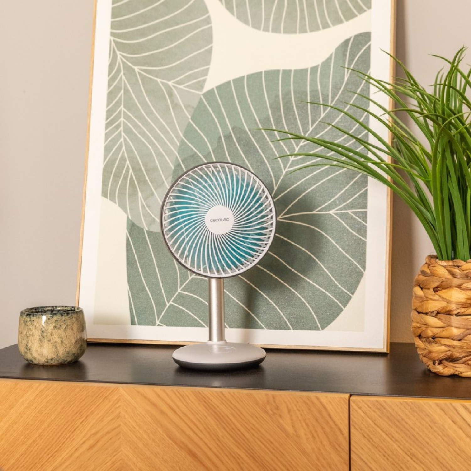 Pequeño ventilador decorativo sobre un soporte, sobre una superficie de madera. Un cuadro y una planta en el fondo.
