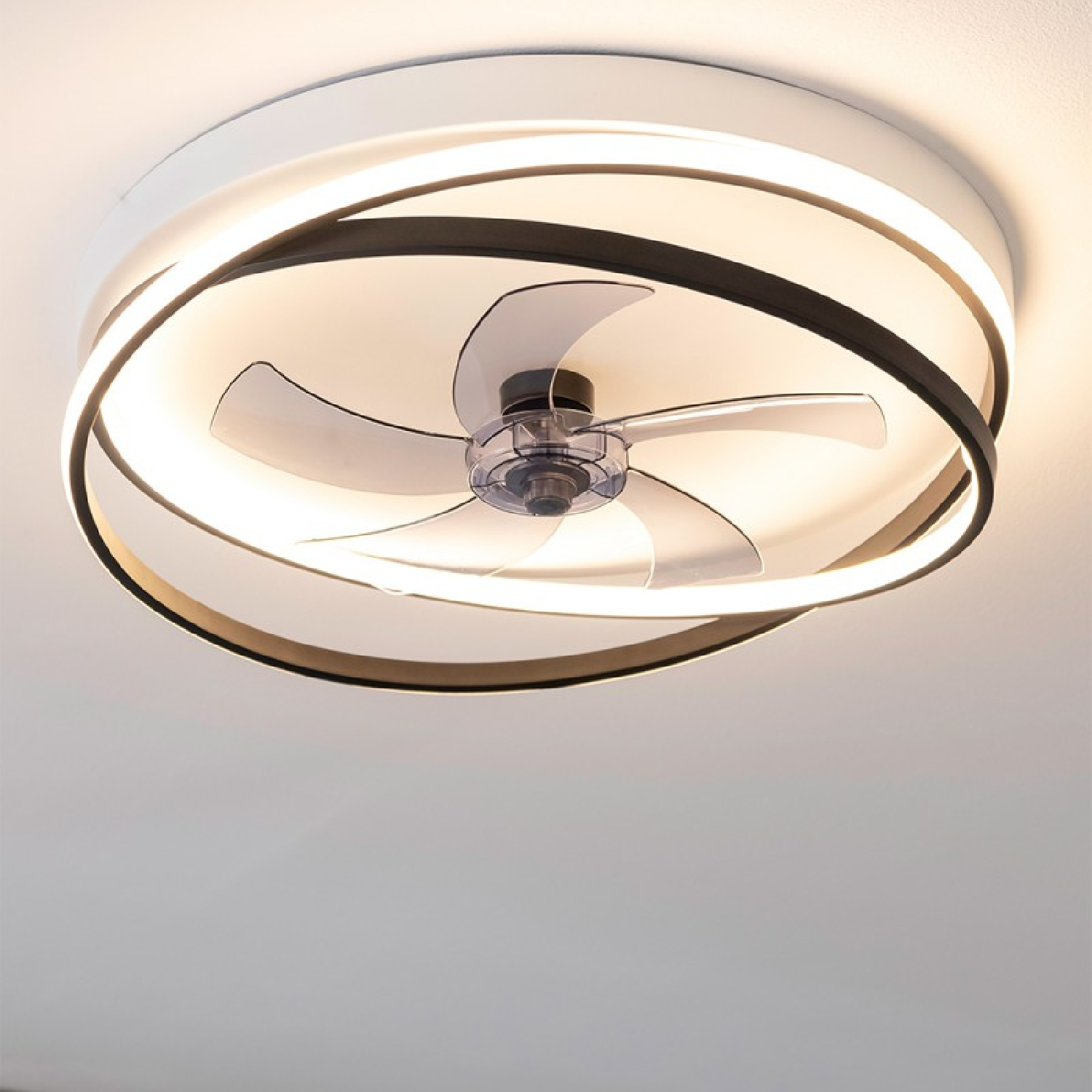 Ventilatore da soffitto con un'apparecchio di illuminazione circolare. Il ventilatore ha tre pale trasparenti. Soffitto bianco con luci accese.