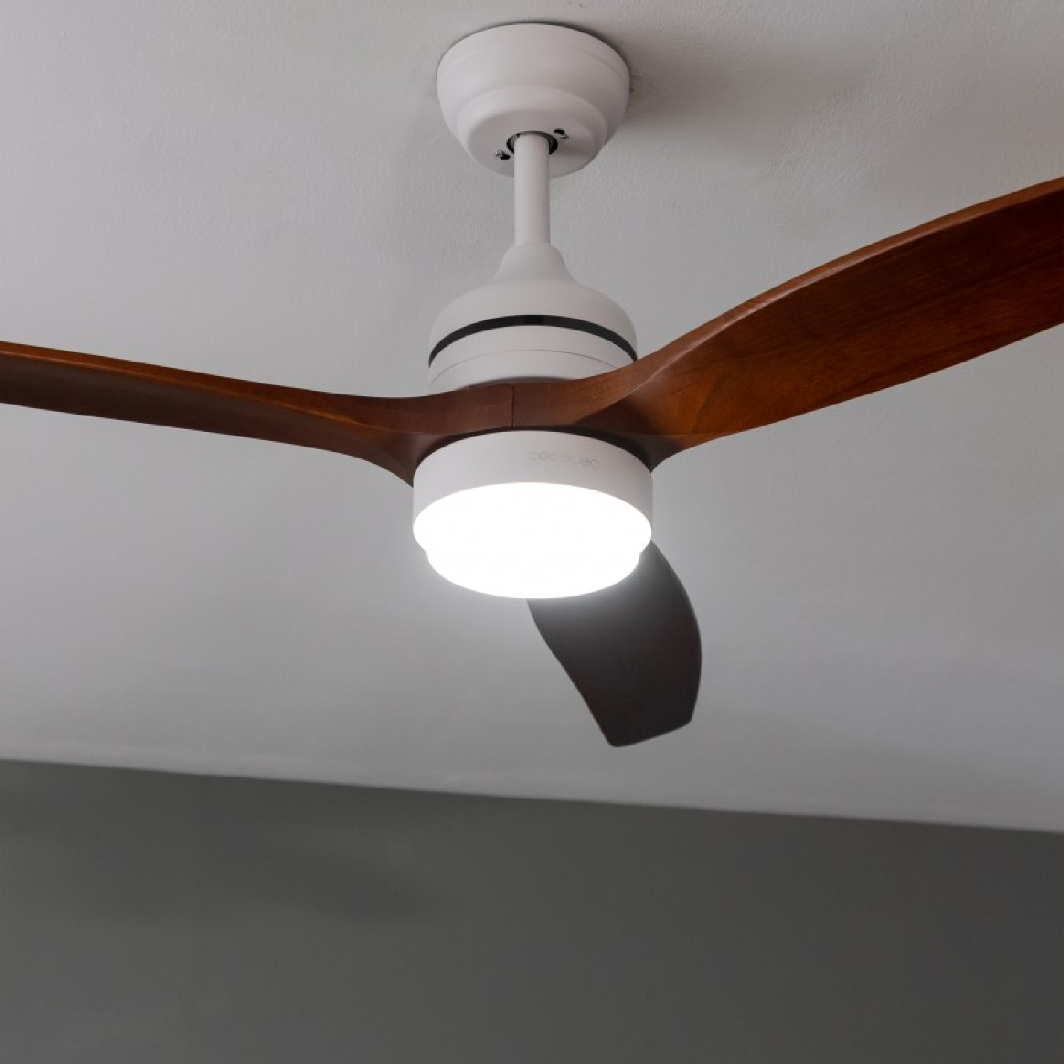 Ein Deckenventilator mit drei Holzflügeln und einer zentralen Leuchte. Der Lüfter befindet sich in einem Raum.