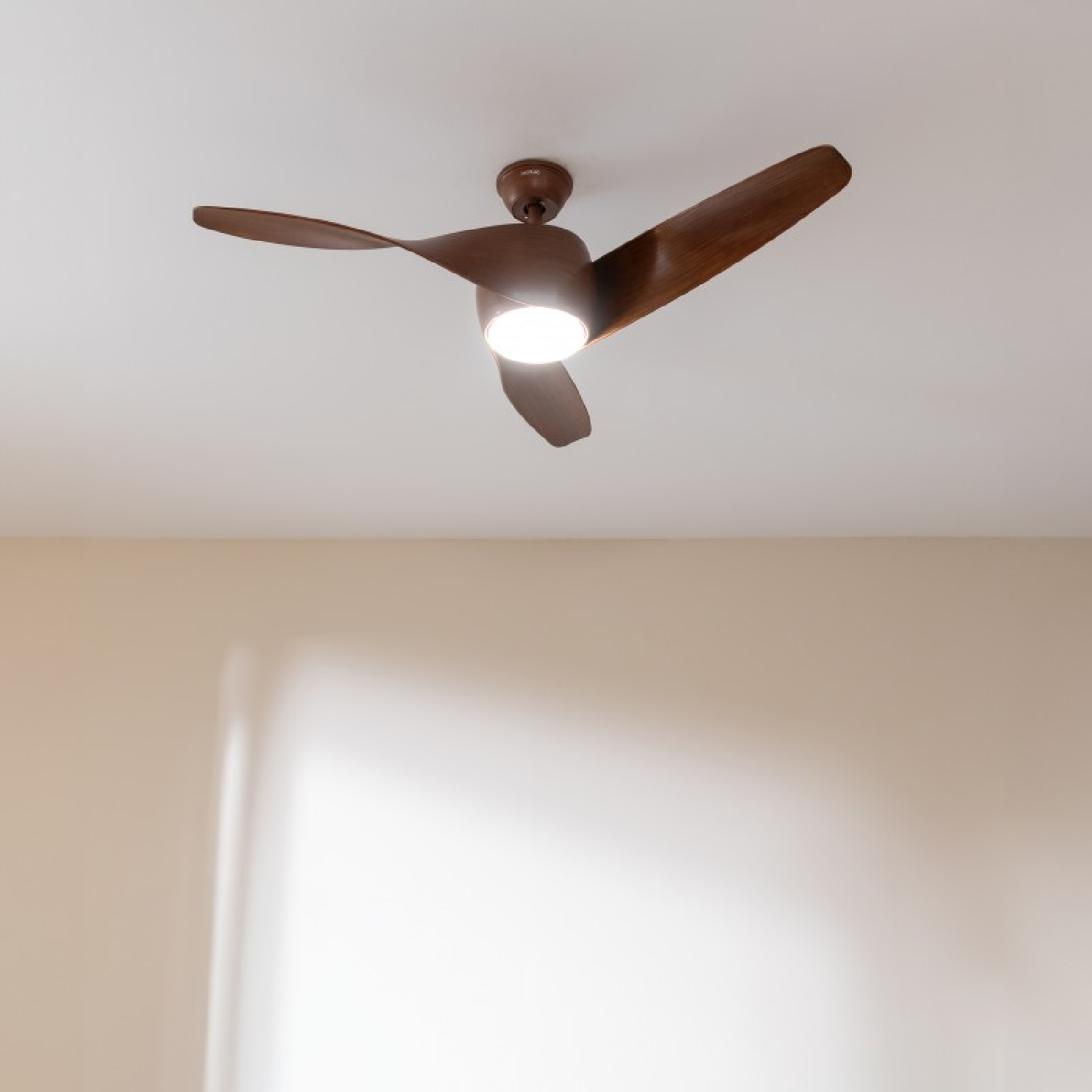 Ventilador de techo marrón con tres aspas, techo blanco y una lámpara en el centro.